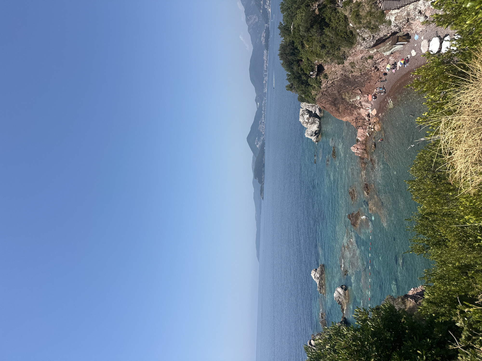 The image captures a vibrant coastal scene in Sveti Stefan, Montenegro, under a clear blue sky. In the foreground, lush green foliage and dry grasses frame a view of the turquoise Adriatic Sea. Rocky outcrops, some submerged and others partially exposed, dot the clear waters. A small, pebbly beach curves along the coastline, populated by a few individuals relaxing under colorful umbrellas and some swimming or wading in the shallows. A wooden walkway with stairs ascends the vegetated cliffside, suggesting access to and from the beach. In the distance, a hazy coastline with rolling hills and buildings stretches across the horizon, hinting at a developed coastal town or city. The overall atmosphere is one of serene summer leisure.