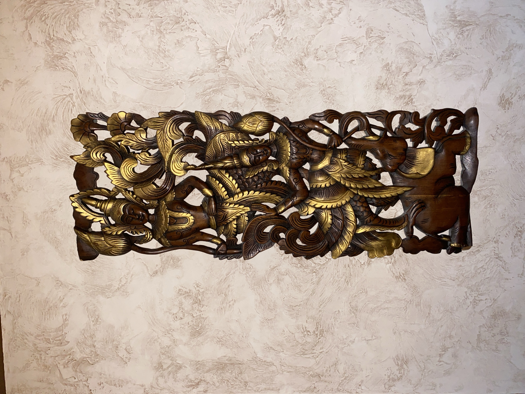 This is a horizontal, close-up photograph of an intricately carved wooden relief panel mounted on a textured, off-white wall. The relief depicts a dynamic scene featuring figures and mythical creatures. The carving is detailed, with sections highlighted in gold paint, creating a striking contrast against the dark wood.
The main subject of the carving appears to be a group of figures engaged in some form of action or combat. One central figure, possibly a warrior or deity, is depicted with a headdress and elaborate wings, suggesting a celestial or powerful being. This figure is intertwined with other forms that could represent animals, possibly a dragon or a mythical bird, also adorned with gold accents. The carving is dense and layered, with flowing lines and organic shapes that convey movement and energy.
Notable details include the fine craftsmanship of the carving, evident in the delicate rendering of feathers, hair, and facial features. The gold paint highlights key elements, drawing the viewer's eye to the most important parts of the composition. The background wall has a textured, stucco-like finish with visible brush strokes or trowel marks, creating a subtle, almost cloud-like pattern that complements the intricate details of the wooden relief without overpowering it.
The lighting appears to be artificial, casting a soft glow on the relief and creating subtle shadows that add depth to the carving. There is no visible text in the image. The overall impression is one of a decorative piece of art, likely from a Southeast Asian culture, given the style of the carving and the depiction of figures and mythical creatures. The setting is domestic, suggesting the relief is hung as a wall decoration. The mood evoked is one of ancient storytelling, mythical power, and artistic mastery.