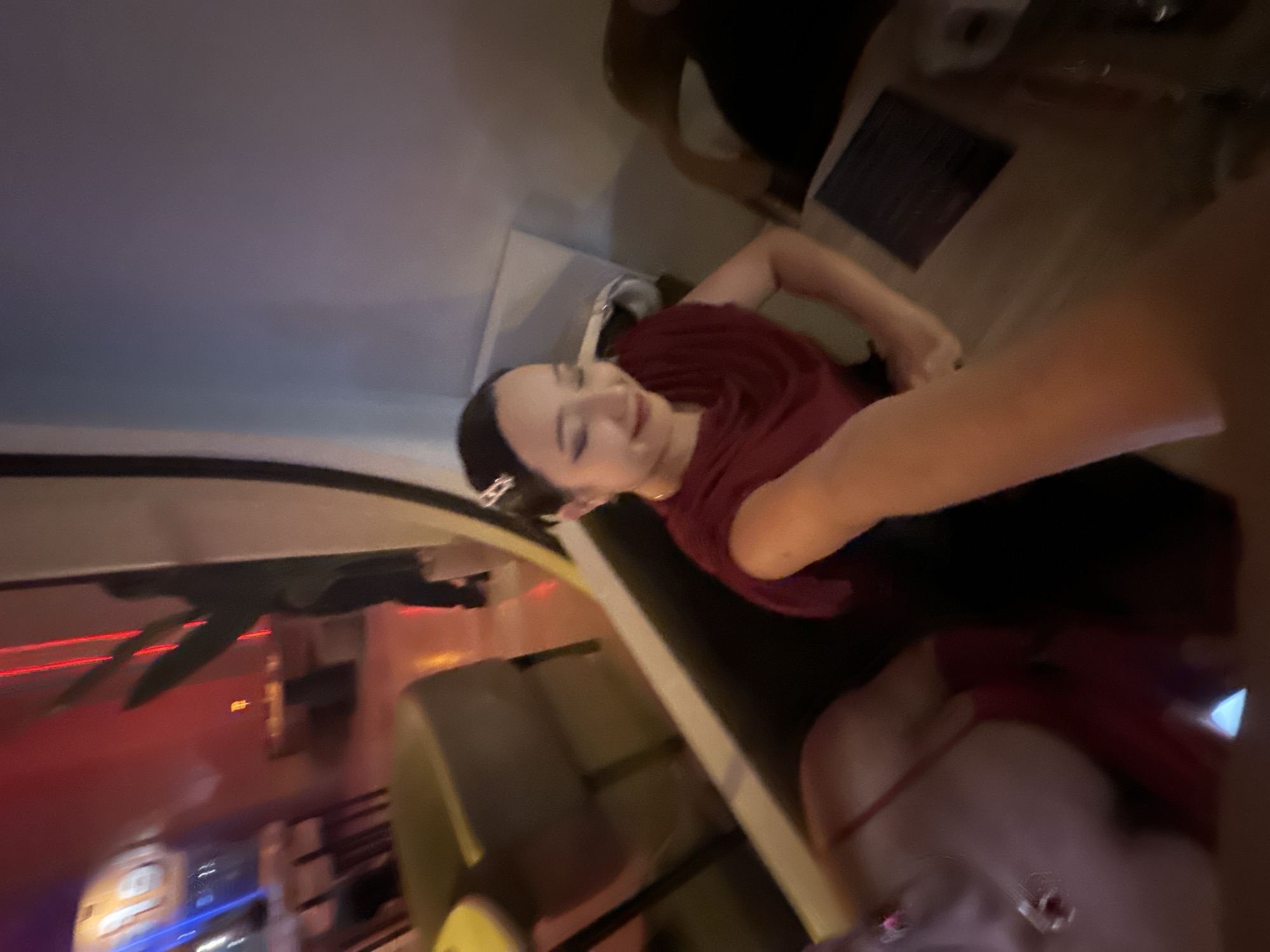 A medium shot shows a person in a maroon shirt seated at a table in an indoor setting, possibly a restaurant or bar. There is a menu on the table. A second person, wearing a red garment and jewelry, is partially visible in the bottom right corner of the frame. A window frames the scene with what may be city lights in the background. Although the city is stated to be North Kuta, Indonesia, visual markers confirming this location are absent.