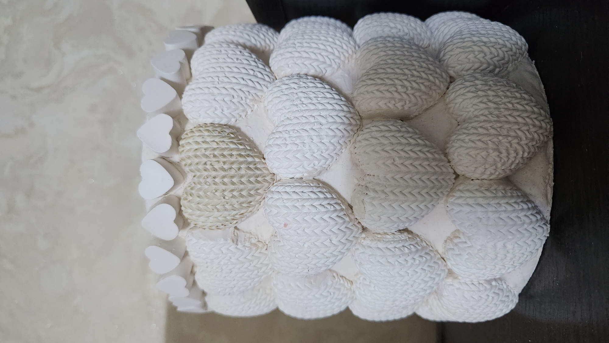 A white, heart-shaped ceramic planter is displayed on a dark surface. The planter is decorated with multiple rows of embossed hearts. The top row consists of small, smooth white hearts. Below this, larger hearts with a textured pattern resembling knitted fabric cover the body of the planter. One heart in the second row from the top appears to be a different shade of off-white or cream and has a slightly rougher texture. The planter is positioned in front of a wall with a light-colored, textured finish. The image does not contain any information about the city of Buynaksk, Russia, nor does it depict any people or actions.