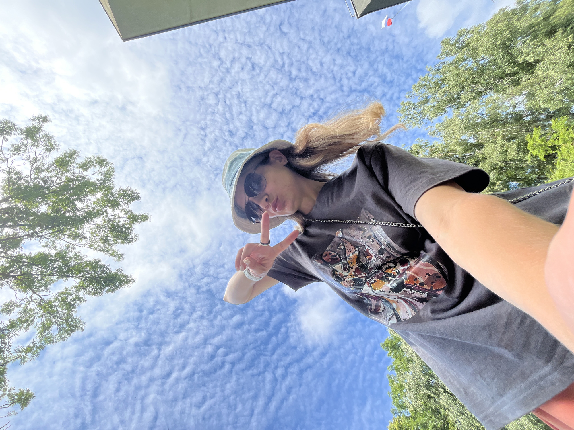 A low-angle selfie shows a young woman wearing a dark t-shirt with a graphic print, a light-colored bucket hat, and sunglasses. She is making a peace sign with her right hand and puckering her lips. Her blonde hair is visible, pulled back into pigtails. The background consists of a bright blue sky with scattered clouds, green trees, and a building corner. A small flag, appearing to be white, red, and blue, is visible in the distance on the right side of the frame. The overall impression is a casual outdoor scene, possibly in a park or open area in the city of Pionersky, Russia.