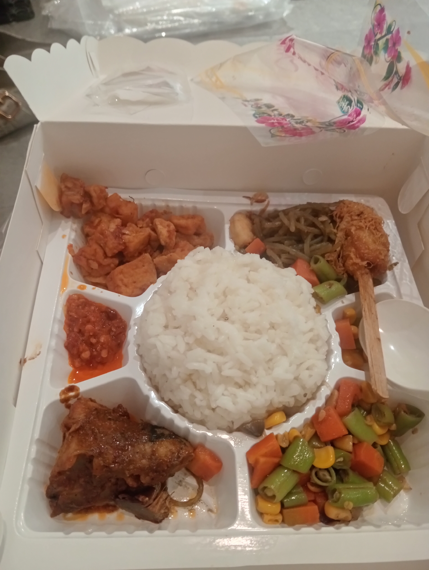 A takeaway food box is positioned on a flat surface. The box is white and divided into several compartments. One compartment contains a mound of white rice. Another holds a piece of fried fish with a dark brown glaze. Adjacent to the rice is a section with what appears to be fried chicken pieces in a red sauce. To the left of the chicken is a small portion of red chili paste. Above the chicken and rice, a compartment contains stir-fried vegetables, possibly green beans and carrots, with some shredded fried material. A wooden skewer with a breaded and fried item, perhaps chicken or fish, is placed in the vegetable compartment. Another compartment contains a mixture of chopped carrots, green beans, and corn. A small white dish is visible in the upper right corner of the box. The lid of the box is open and displays a floral pattern. The scene is well-lit, with the food appearing to be freshly prepared. The image was taken from a slightly elevated angle, looking down into the food box. The location is specified as Larangan, Indonesia.