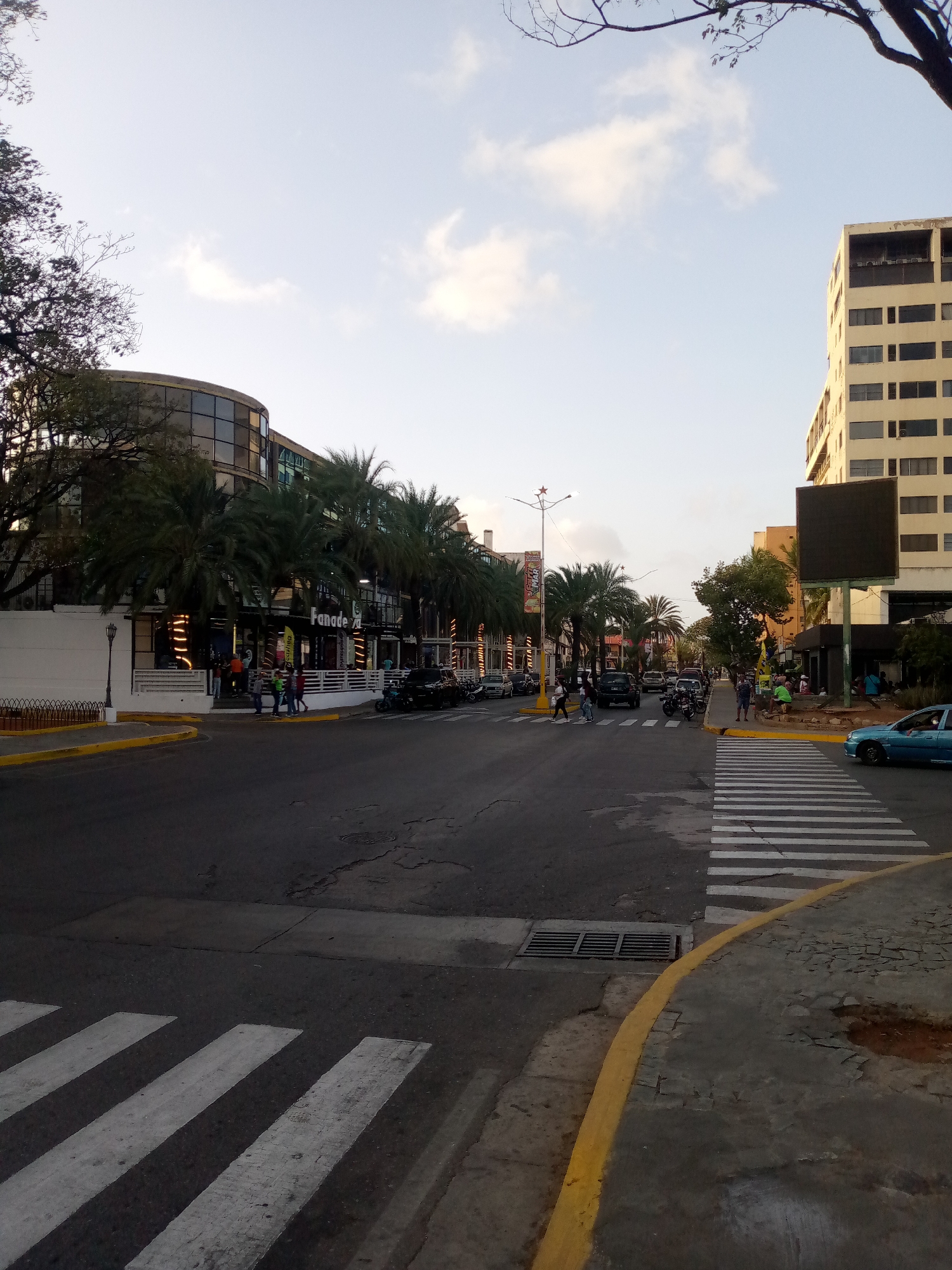 The image depicts a street scene in Juan Griego, Venezuela. A multi-lane street with a crosswalk is visible, with traffic including cars, motorcycles, and pedestrians crossing. The street is lined with palm trees and buildings. A multi-story office building is on the right of the street. On the left is a curved-facade, glass-enclosed building. It appears to be a restaurant or commercial establishment (sign says "Fanade..."). Yellow painted curbs border the streets. Overhead, a cloudy sky is visible.