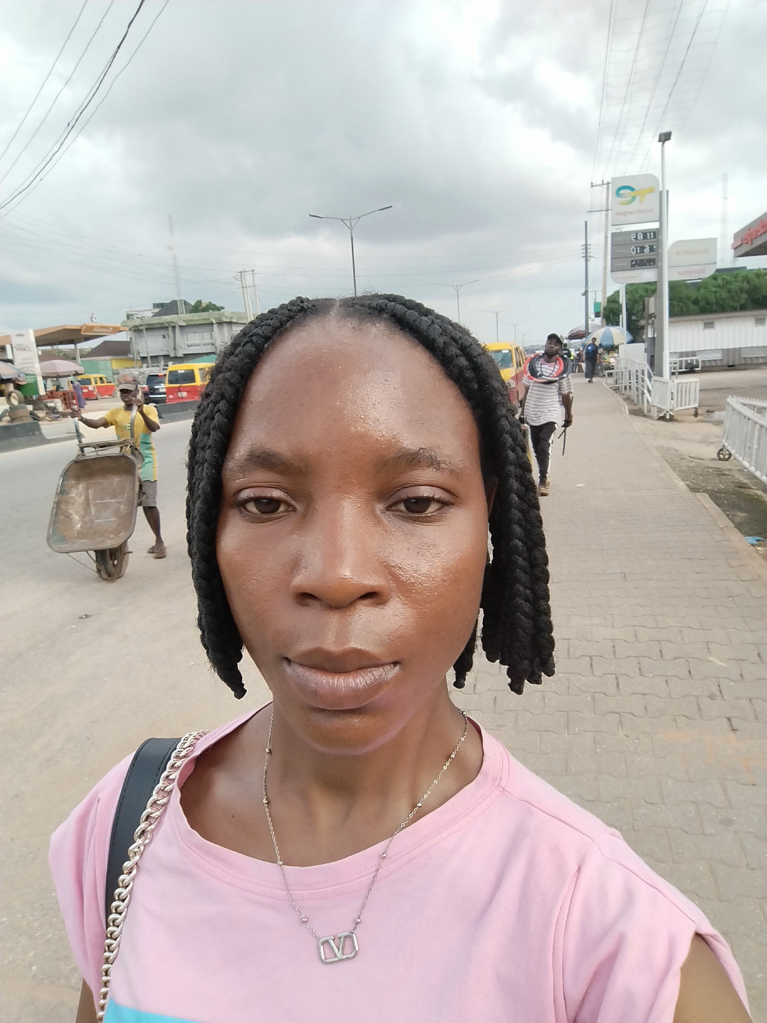The image shows a selfie taken outdoors on a cloudy day in Benin City, Nigeria. The foreground features a woman with her face and upper body visible, wearing a pink t-shirt and a silver necklace with a "V" pendant. Her hair is styled in two prominent braids.
In the background, there are several elements indicating an urban environment. A man is pushing a wheelbarrow on a paved surface that appears to be a road or a large walkway. He is wearing a yellow shirt and grey shorts. Further back, several vehicles, including yellow minibuses, are visible, suggesting a street scene with traffic. A gas station sign with prices displayed is also present, identifiable by the "ST" logo. In the distance, buildings and power lines can be seen, typical of a city landscape. A man in a striped shirt is walking away from the camera along the paved walkway. The overall scene depicts a busy street in Benin City.