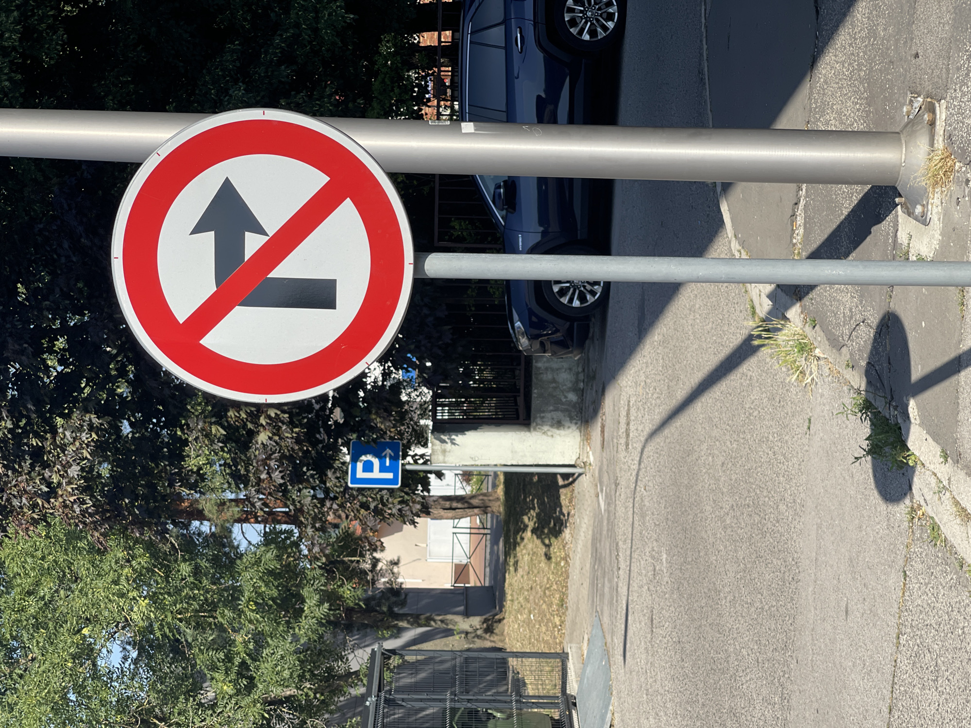 The image shows a traffic sign indicating "no right turn" and "no u-turn" in a residential area. The sign is circular with a red border and a diagonal red line across it. Inside the circle, there is a black arrow pointing upwards and a black arrow pointing to the right, with the diagonal line crossing both.
The sign is mounted on a metal pole, which also supports another metal pipe. To the right of the sign, a dark blue car is parked. In the background, there are trees, buildings, and a paved road. A blue parking sign with a white arrow pointing upwards is visible to the left of the main traffic sign.
The scene appears to be outdoors during the daytime, with clear weather. The sun casts shadows on the ground. The location is not explicitly identified, but the presence of trees, buildings, and a road suggests an urban or suburban environment. No people are visible in the image. The overall mood is neutral, depicting a typical street scene with traffic regulations.