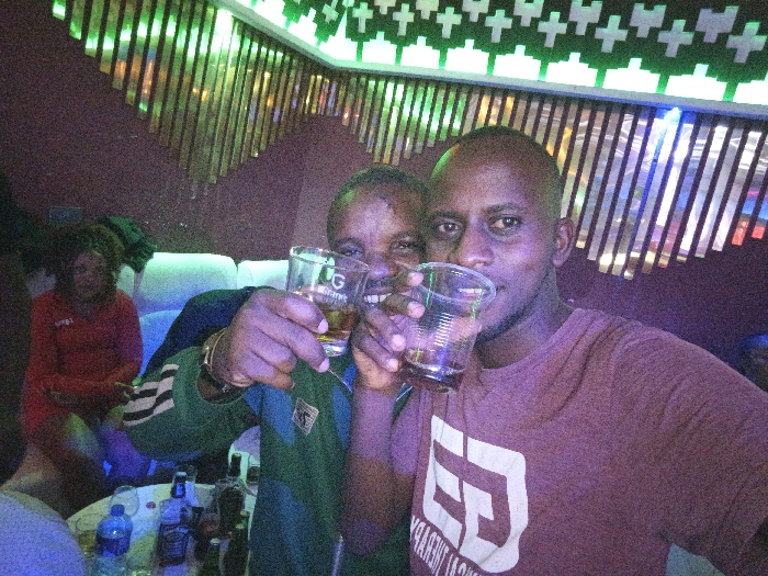 Two men raise plastic cups with liquor, grinning at the camera in a dimly lit bar. Behind them, a woman in a red outfit sits on a white chair. The bar is decorated with lights, and bottles sit on a table in the foreground. The men are close together, posing for the photo. One wears a green jacket, and the other a maroon t-shirt with a white logo.