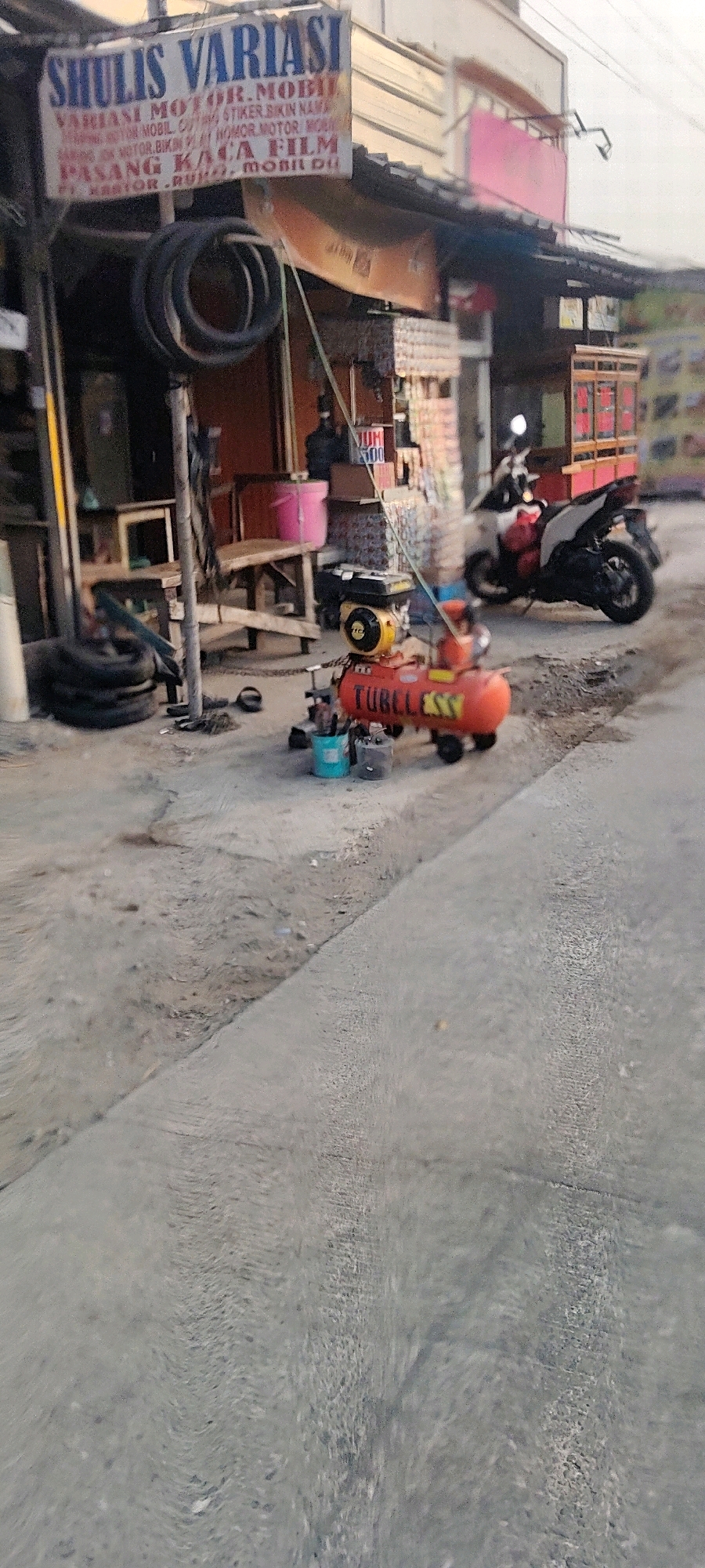 The image captures a street scene in Mekarmukti, Indonesia, focusing on a small shop specializing in vehicle modifications and accessories. A prominent sign, "SHULIS VARIASI," in blue and red lettering, hangs above the storefront, detailing services like "VARIASI MOTOR. MOBIL," "STIKER," and "PASANG KACA FILM."
In front of the shop, various items are visible. A red air compressor with the brand "TUBELESS" is positioned on the ground, alongside two buckets, one blue and one dark-colored. Several tires are stacked near a wooden bench and some shelves displaying merchandise, including what appear to be small boxes of items. A motorcycle is parked to the right of the air compressor, and further back, another parked motorcycle is partially visible. A person, likely a customer, is seated on a stool inside the shop, partially obscured. The ground is a mix of asphalt and dirt. The overall scene suggests a bustling, everyday operation of a local business in Indonesia.