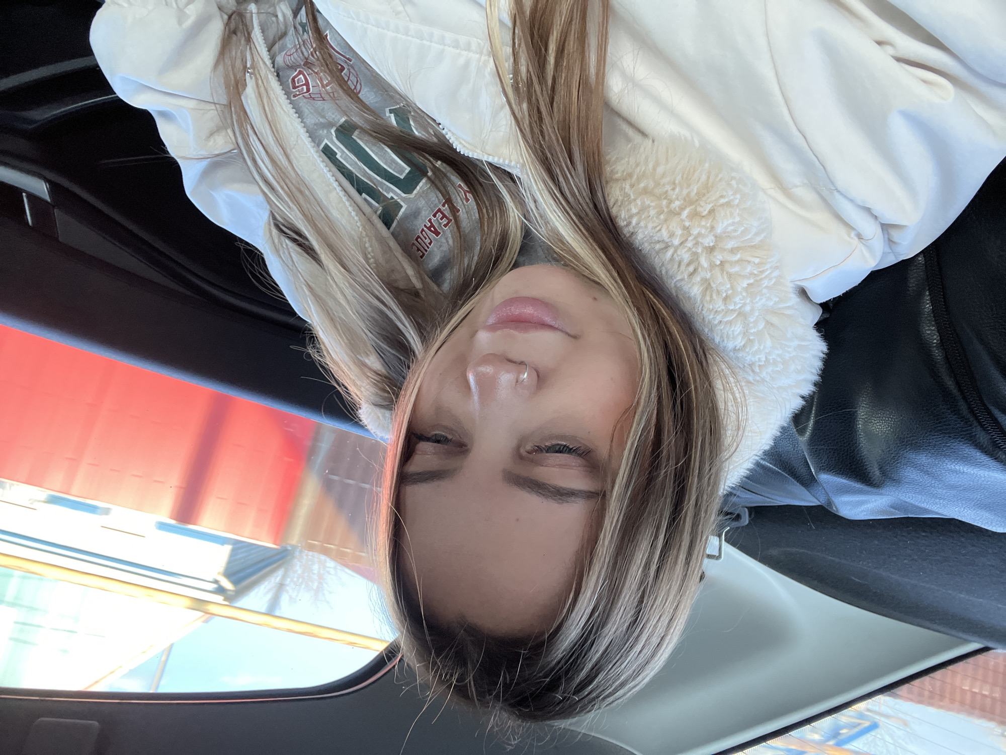 A young woman with long, highlighted hair is seated in what appears to be a car. She is wearing a gray t-shirt with green lettering under a white jacket that has a fluffy, light-colored trim. Her face is angled towards the camera, with closed eyes and visible nose ring. Outside the car window, a building with red and white elements is visible, suggesting the location may be in an urban area. The sky is visible in the reflection on the car window, appearing clear and bright. The interior of the car includes black leather components, potentially part of the seat or dashboard. The photograph is taken at an odd angle, rotated such that the woman appears upside down. The location is Novopokrovskaya, Russia.