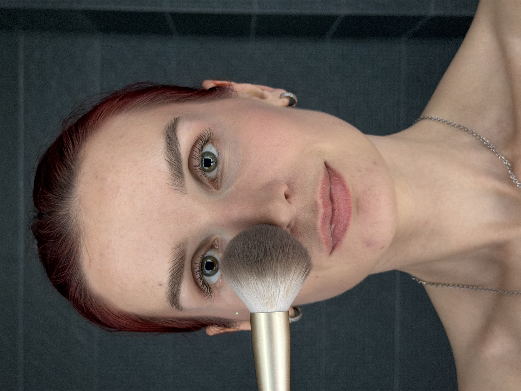 A close-up, sideways portrait shows a person, likely female, with reddish-brown hair and fair skin, set against a dark, horizontally tiled background. The person is holding a large, fluffy makeup brush with a gold handle and multi-toned bristles (brown fading to white) near their nose and cheek, suggesting the application of makeup.
The individual has striking green or hazel eyes with visible eyelashes and well-defined eyebrows. Their expression is neutral and direct, with a slight hint of a smile. Lips are lightly colored with what appears to be lipstick or gloss. Notable features include a silver hoop earring in the left ear and a thin silver chain necklace visible around the right shoulder/neck area. The skin shows natural texture and minor imperfections. The lighting is soft, emphasizing the facial features and the texture of the makeup brush. The overall composition is a detailed, intimate shot of a beauty routine or portrait. No text is visible in the image. The location context for this image is Chaikovsky, Russia.