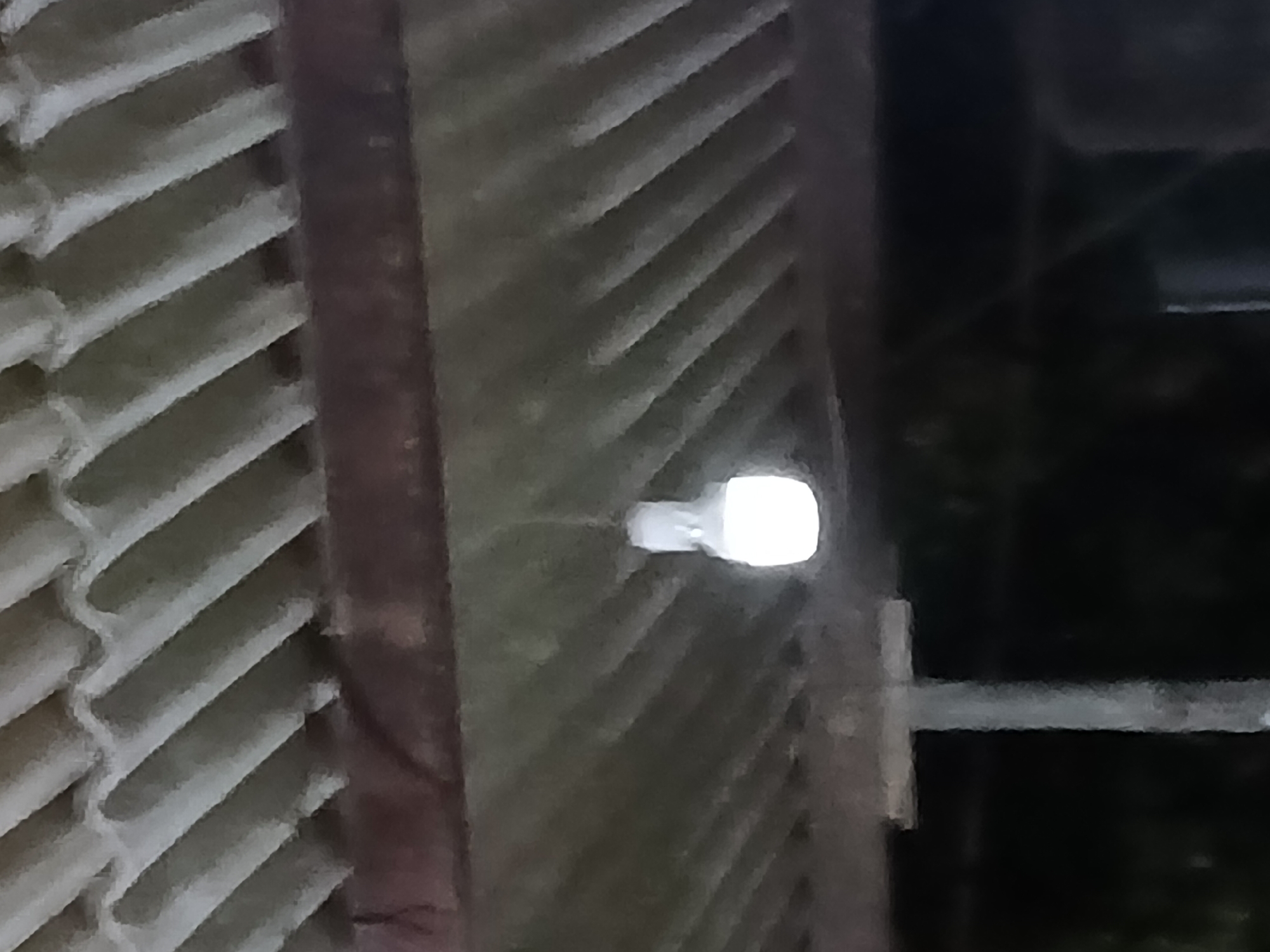 An illuminated LED light bulb hangs from a wire suspended from a corrugated metal roof, casting a soft glow on the understructure of the roof. The roof is supported by a metal beam and a vertical pole, which are visible in the foreground. The scene appears to be an outdoor setting, possibly a porch or shelter, in Altagracia, Venezuela. The lighting suggests it is either dusk or night. The image is grainy, indicating it was taken in low light conditions.