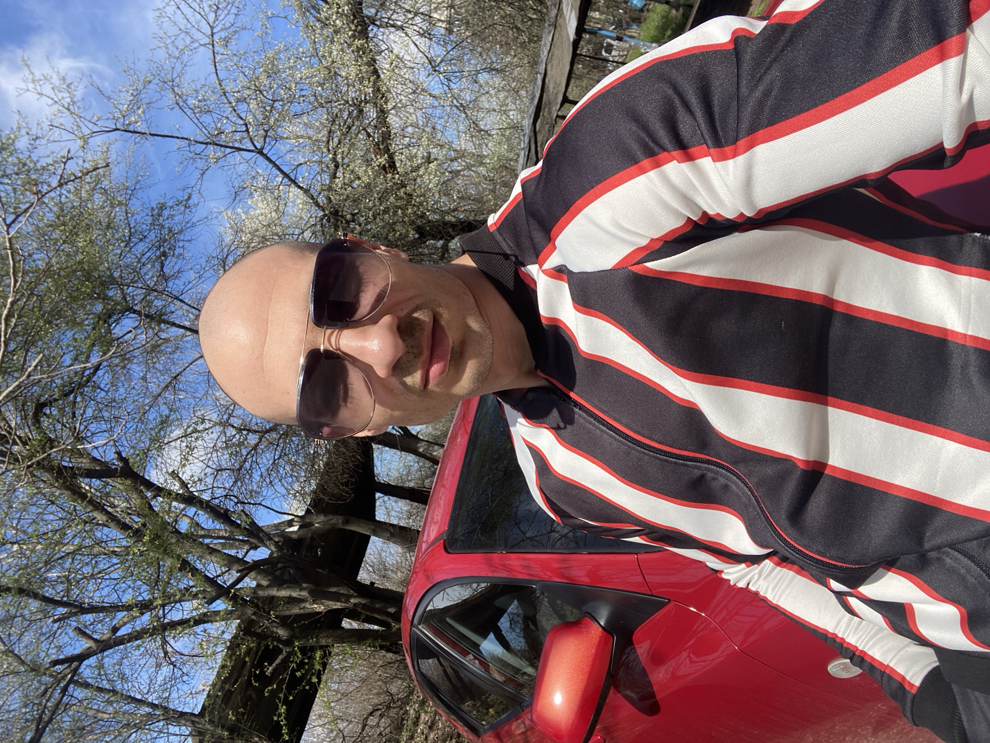A bald man with a mustache, wearing sunglasses and a striped black, white, and red jacket, poses beside a red car. The scene is outdoors, under trees with budding foliage and a blue sky visible through the branches. The location is Pleven, Bulgaria.