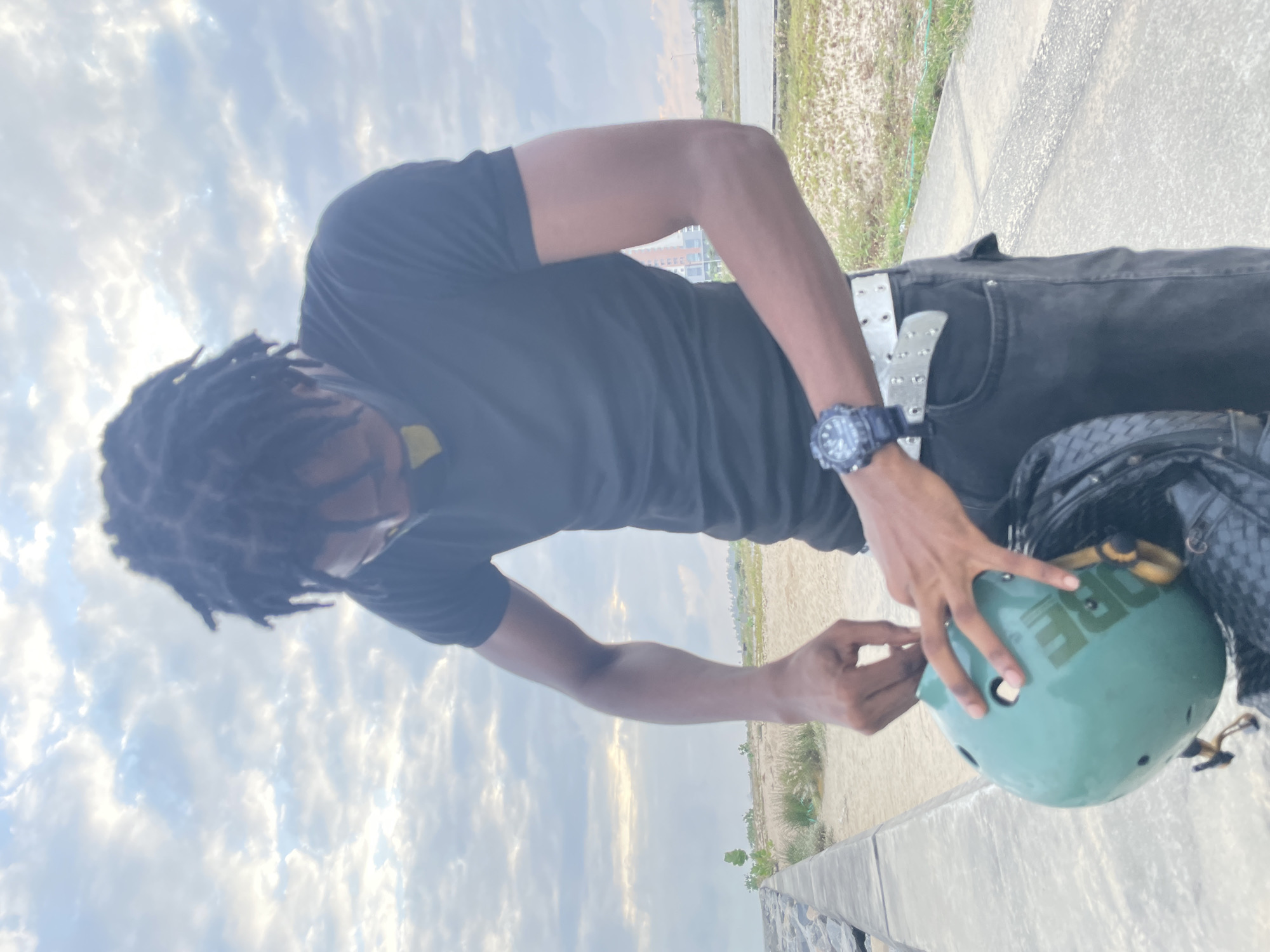 An adult male with dreadlocks wearing a black t-shirt and dark pants is shown from the waist up outdoors. He is holding a teal-colored helmet with yellow straps and appears to be adjusting it. A black backpack is visible below the helmet. He is wearing a digital watch on his left wrist and a white studded belt. The background shows a cloudy sky, a sandy area, and some distant buildings. This scene is located in Inupa Kekere, Nigeria.