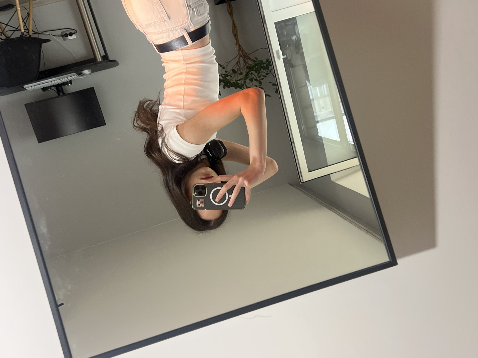 This media file presents a mirror selfie of a young person, depicted upside down, likely taken in a room in Ternopil, Ukraine. The central subject is a person with long, dark hair, wearing a white short-sleeved t-shirt and light-colored pants with a dark belt. They are holding a smartphone, which has a dark case with a circular light-colored design, pointed towards the mirror to capture their reflection. Headphones are visible draped around their neck.
The background, visible in the mirror's reflection, shows elements of a room. To the viewer's left (upper right in the actual scene), there is a black shelf or desk holding a black pot, various wires, a white computer keyboard, and a black computer monitor. To the viewer's right (upper left in the actual scene), a window with a white frame is visible, reflecting a building exterior, suggesting a daytime setting with ample natural light. A plant with green leaves and branches can be seen near the window. A reddish tint is visible on the person's right arm, possibly from light reflection. The image appears to be rotated 180 degrees from its original capture. No specific readable text is discernible on the phone or other objects, apart from the design on the phone case.