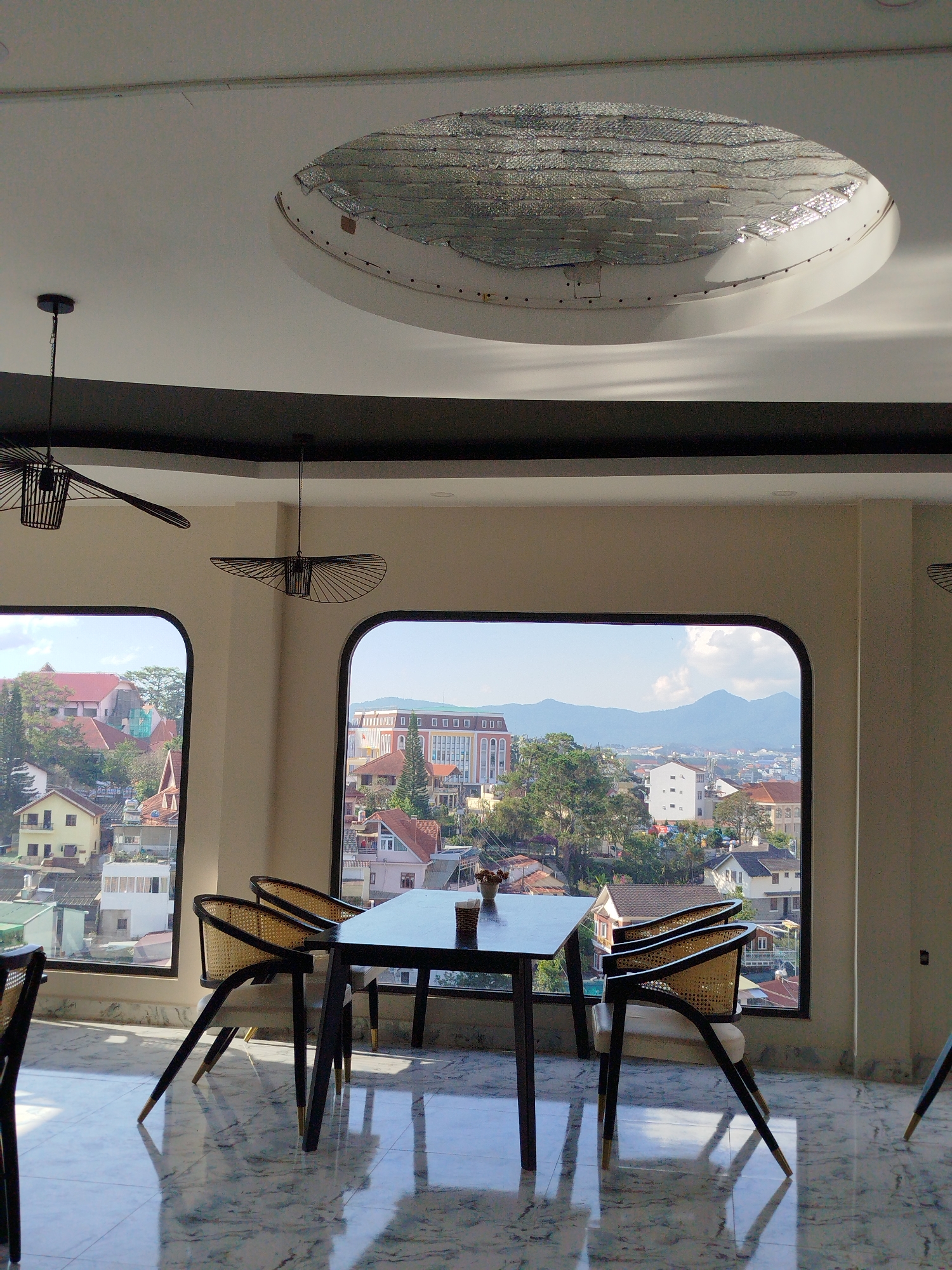 The image captures a bright, upscale dining space in Da Lat, Vietnam, with a view overlooking the cityscape. A black square table is centered between two large, rounded windows, surrounded by four chairs with black frames and woven backs. The chairs' legs are angled and have gold accents at the bottom. On the table sits a small arrangement, possibly a flower vase.
Outside the windows, the scene reveals a vibrant cityscape with a mix of residential and institutional buildings, set against a backdrop of rolling hills and distant mountains under a bright sky.
The interior design includes a glossy, marble-like floor that reflects the light and adds to the room's brightness. Overhead, modern, minimalist chandeliers hang from the ceiling. The ceiling features a decorative, recessed oval panel with a textured, reflective material.