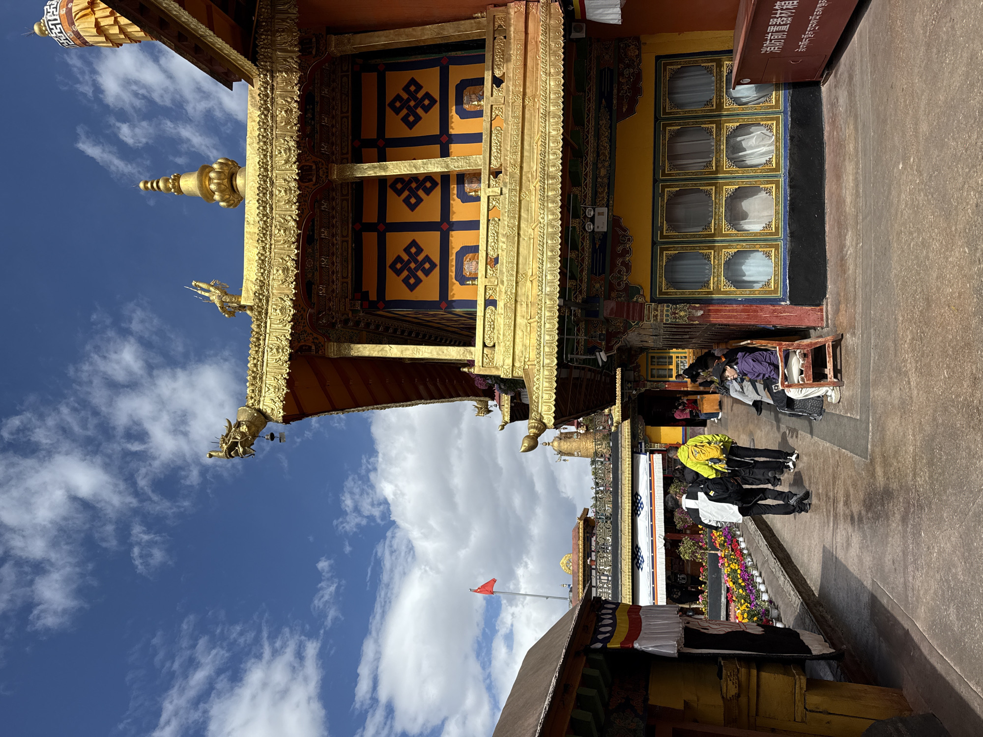 The image shows a building in the city of Chengguan District, China. The building features ornate gold detailing, particularly around its roof and columns. A banner with blue and yellow designs hangs on the building's front. Adjacent to the building is a walkway where several people are present. A row of colorful flowers sits near the walkway's edge. Beyond the building, a red flag is flying, and the sky is visible with scattered clouds.
