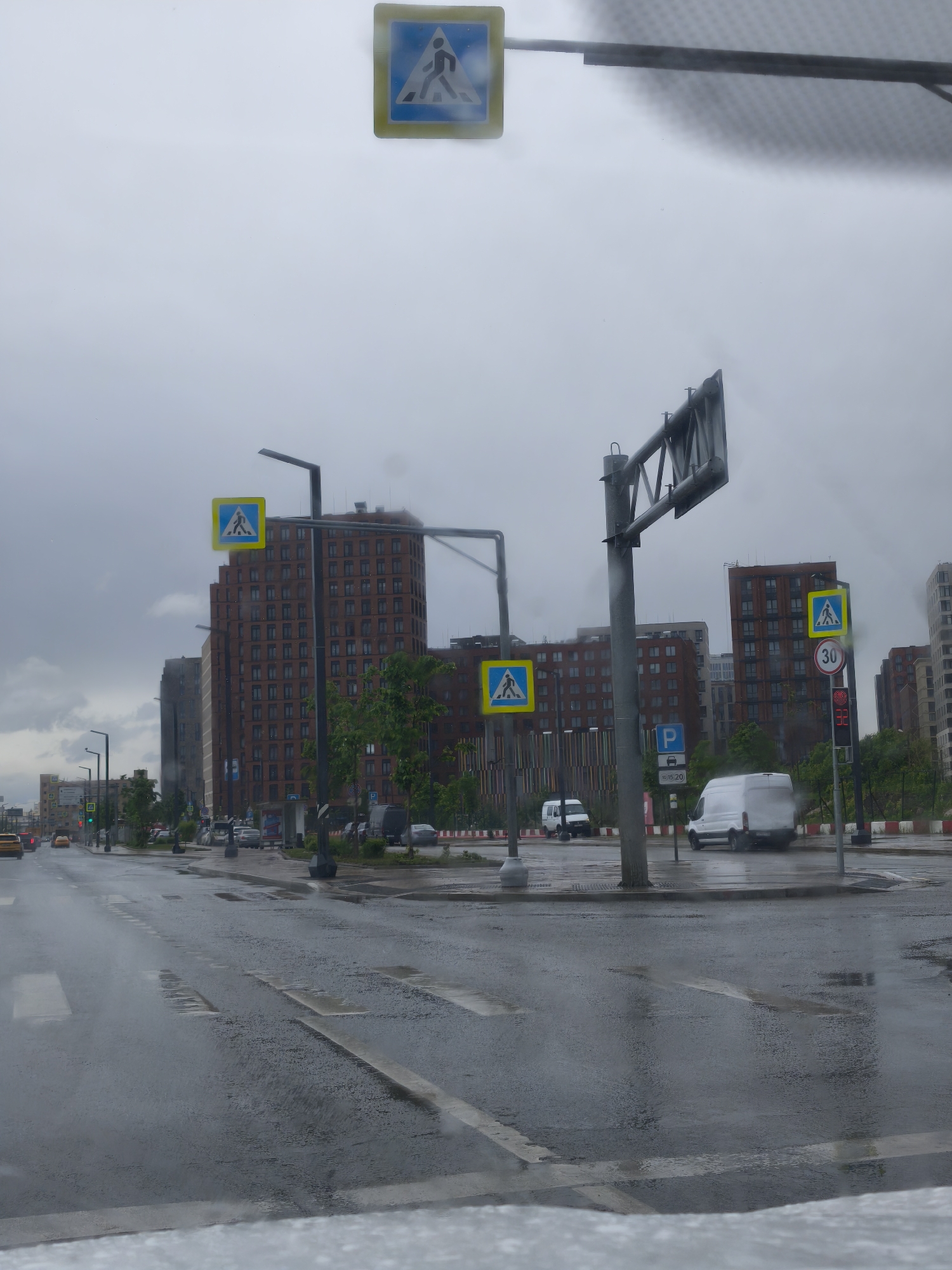 The image depicts a street scene in Moscow, Russia, on a rainy day. The foreground is dominated by a wet road with pedestrian crosswalk markings. Several cars and vans are visible on the road and parked alongside the street. Multiple pedestrian crossing signs are positioned throughout the scene. In the background, there are tall brick buildings. Streetlights and traffic signals are also visible.