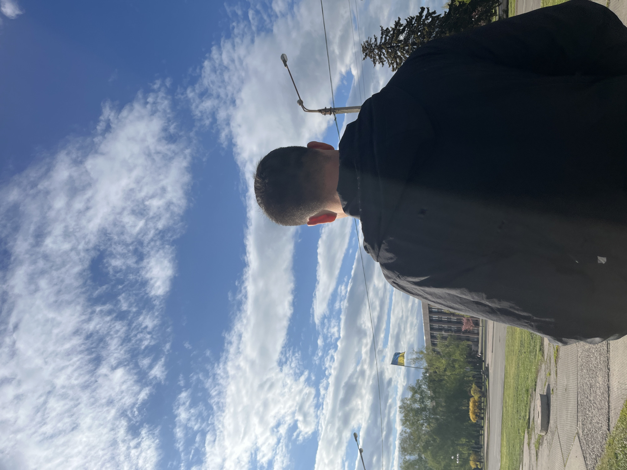 A low-angle shot shows a close-up of a man's head and upper back, wearing a dark jacket, against a backdrop of a cloudy blue sky. The shot appears to be taken on a city street with a glimpse of trees, a building, a street light and the flag of Ukraine. The location is Unknown.