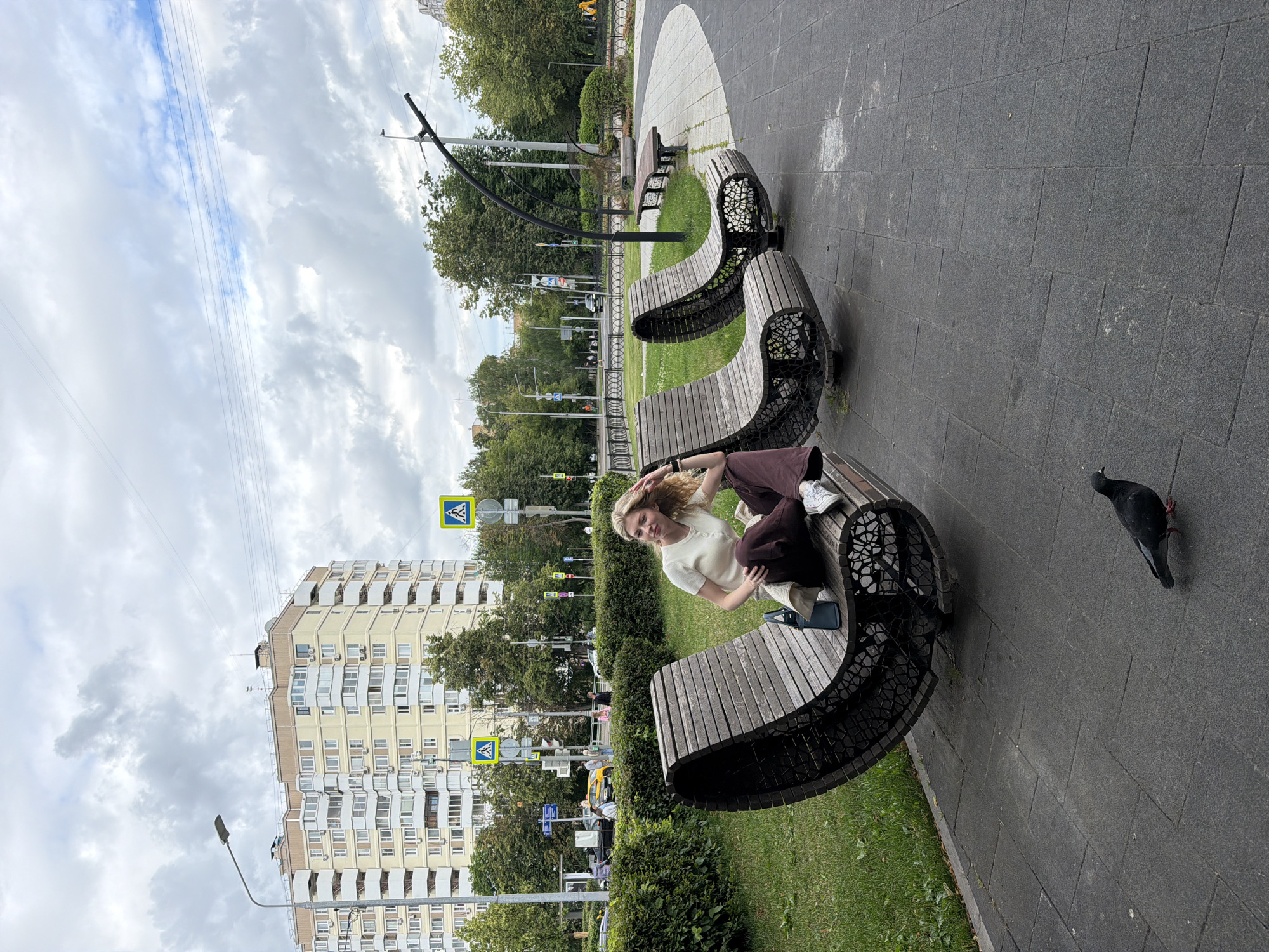 A young woman with blonde hair sits on a curved wooden bench in an urban park. She is wearing a light-colored sweater and dark trousers, and her legs are crossed. She looks towards the camera with a relaxed expression. Beside her on the bench is a dark-colored phone. A pigeon stands on the dark grey paved walkway to her right. The bench is part of a modern seating area with a decorative metal base. Behind the bench, a strip of green grass is bordered by a dark metal fence. Further in the background, trees and buildings are visible, including a tall, light-colored apartment building with many balconies. Several pedestrian crossing signs are scattered throughout the scene. The sky is cloudy and overcast, suggesting it could be daytime. The overall scene depicts a moment of rest in a city park, with elements of nature and urban infrastructure coexisting.