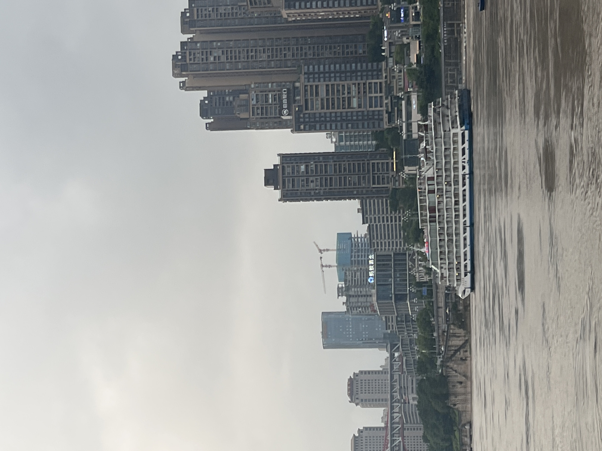 An overcast, daytime view of a wide, murky river in Nan'an District, China, with a modern cityscape on the far bank. The foreground is dominated by the river's surface, which is rippled and a muddy brown color. In the middle ground, a large, white cruise ship with blue accents is sailing from left to right. It has multiple decks with numerous windows. Behind the ship, the riverbank is lined with a retaining wall and some greenery. The background features a dense urban skyline composed of numerous tall, modern buildings of varying architectural styles and heights. Some buildings are under construction, indicated by cranes. The sky is uniformly gray and cloudy, obscuring the horizon.