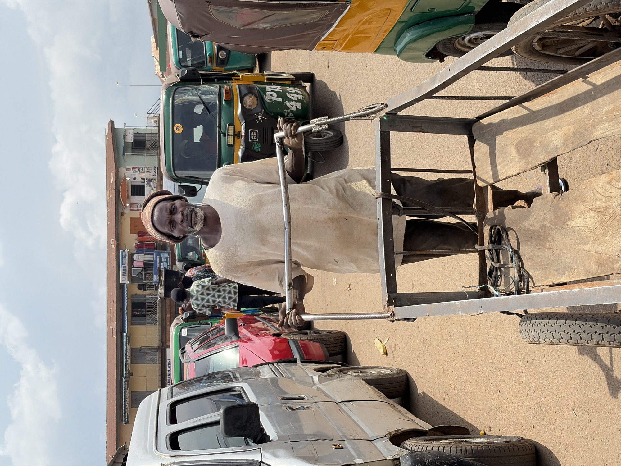 A man wearing a light-colored t-shirt and a beanie pushes a cart in a street in Jos, Nigeria. The man is in the foreground, looking directly at the camera. The cart is made of metal and wood, with two wheels at the back. Behind the man, several auto rickshaws, a red car, and a silver van are parked in the street. Buildings line the street in the background. The sky is cloudy.