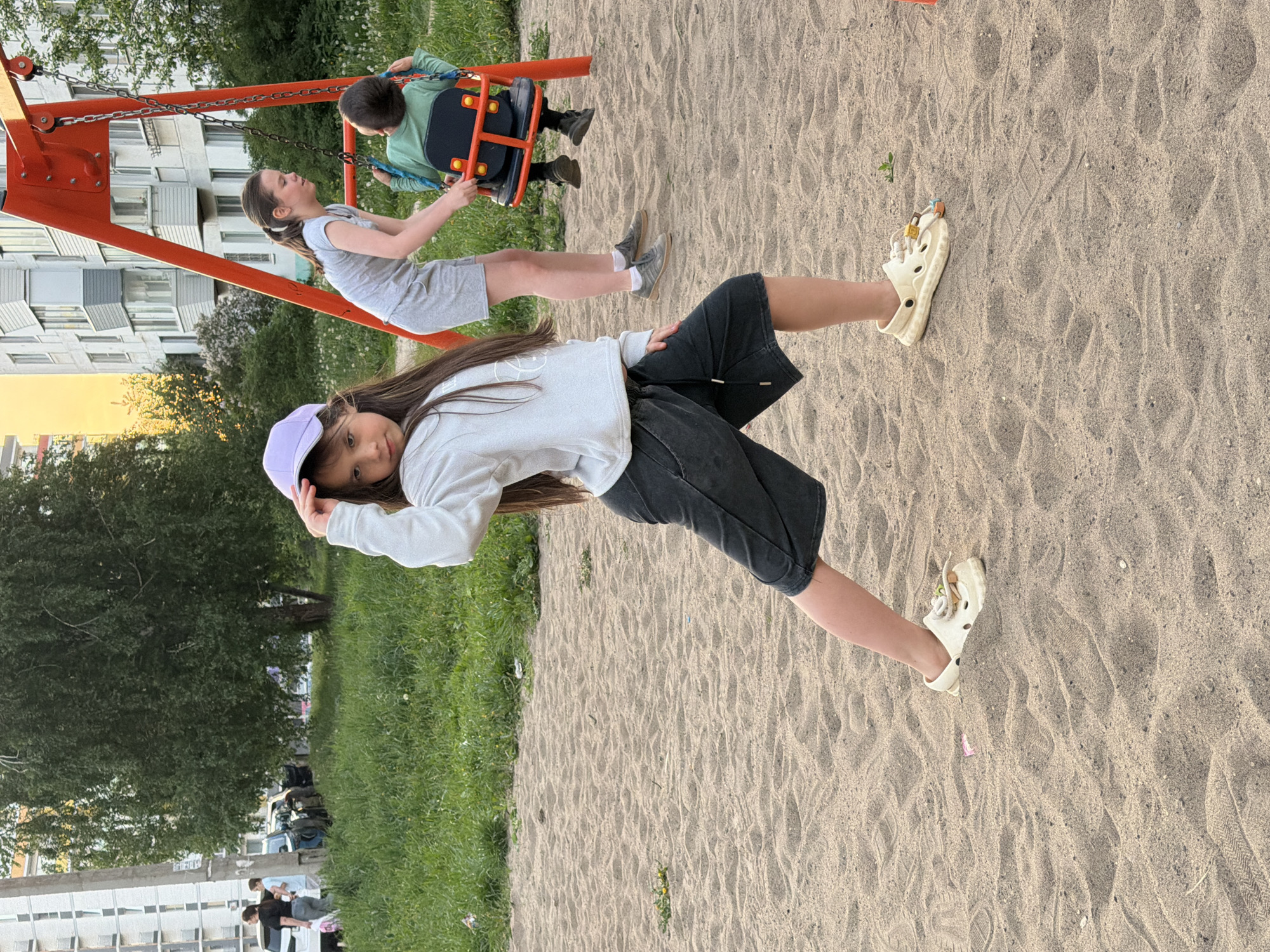 The image shows three children at a playground. A girl with long brown hair, wearing a light purple baseball cap, a white long-sleeved shirt, dark shorts, and beige Crocs stands in the foreground, slightly leaning towards the camera. In the background, two children are on a swing set. One child, wearing a grey dress, is sitting on a regular swing. The other, wearing a green shirt, is on a bucket swing. The playground is covered in sand, with patches of grass on the sides. In the background, a building with many windows is visible. The location is most likely in Chaikovsky, Russia.