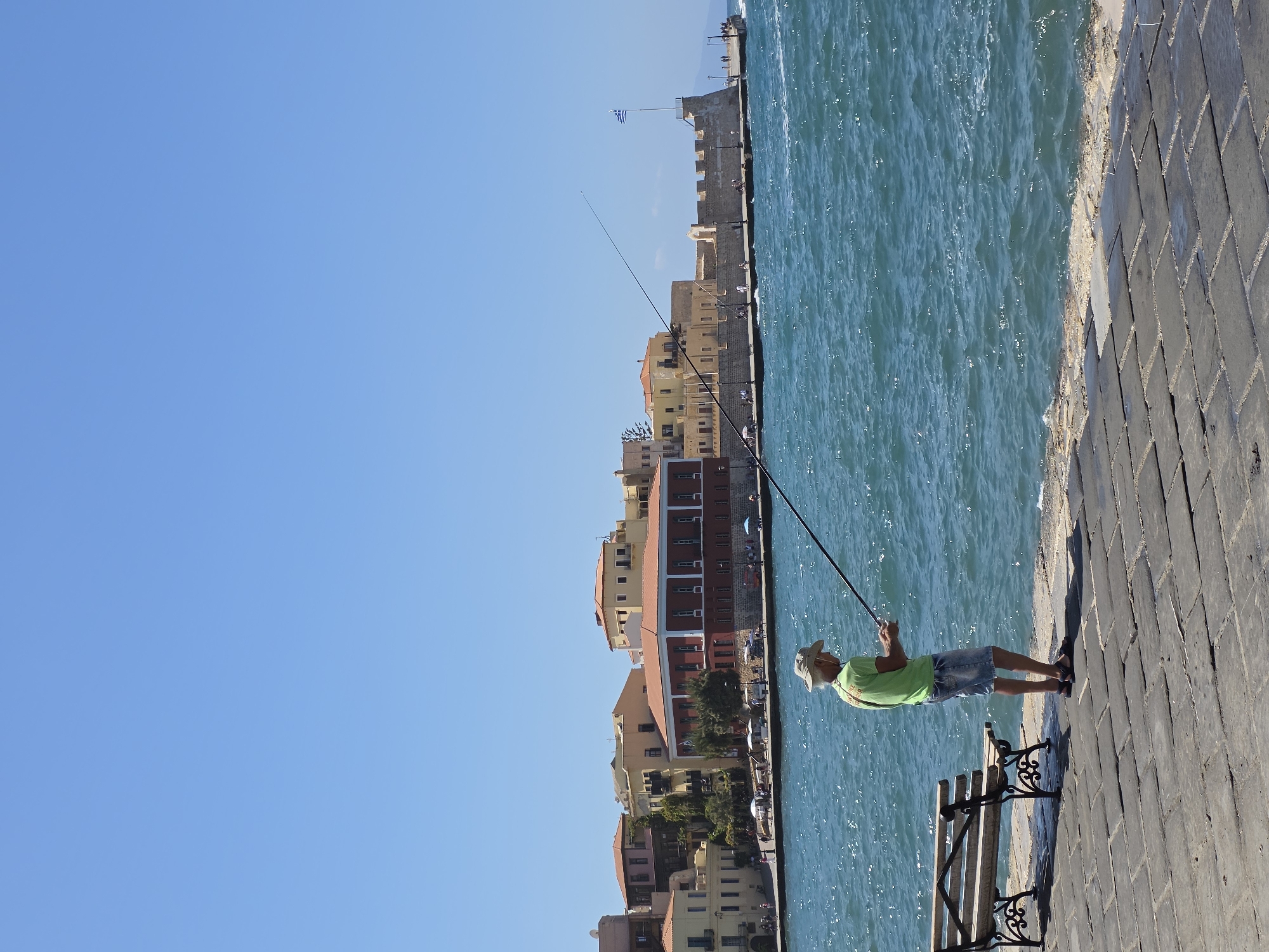 In the city of Chania, Greece, a lone man fishes from a stone promenade. The man, wearing a light-colored hat, a green t-shirt, and denim shorts, stands on the right side of the frame, holding two fishing rods extended over the blue, choppy water. To his left, a wooden bench with ornate black metal legs sits on the promenade. In the background, a row of buildings with red-tiled roofs line the waterfront, varying in color from yellow to peach and red. A Greek flag flies from a stone structure to the right of the buildings. The sky is clear and blue, with no clouds.