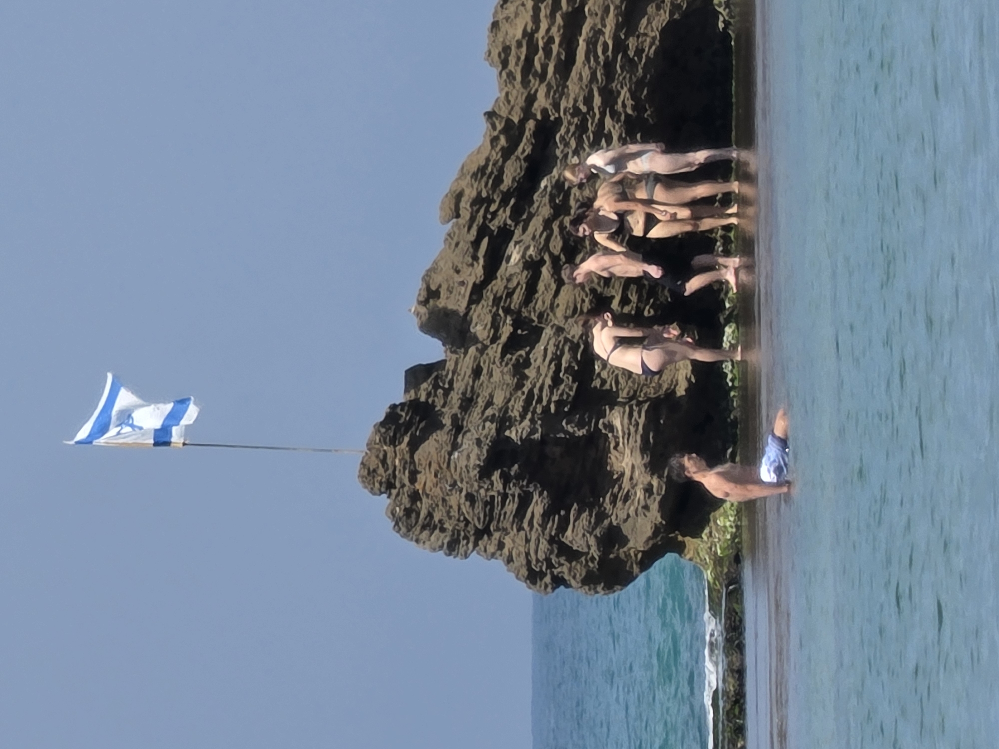 A long, vertical shot shows a group of people on and near a rocky outcrop in the sea at Bat Yam, Israel. From left to right, an Israeli flag is posted on the land, with the blue and white flag clearly visible against the bright, light blue sky. The outcrop itself appears rough and dark, contrasting with the lighter tones of the sky and sea. The group of people stand closely together in a line, and all are partially undressed. In the water, a man swims with a pair of blue and white swim trunks. The sea appears calm and blue, reflecting the light. The overall scene captures a sunny day at the beach.
