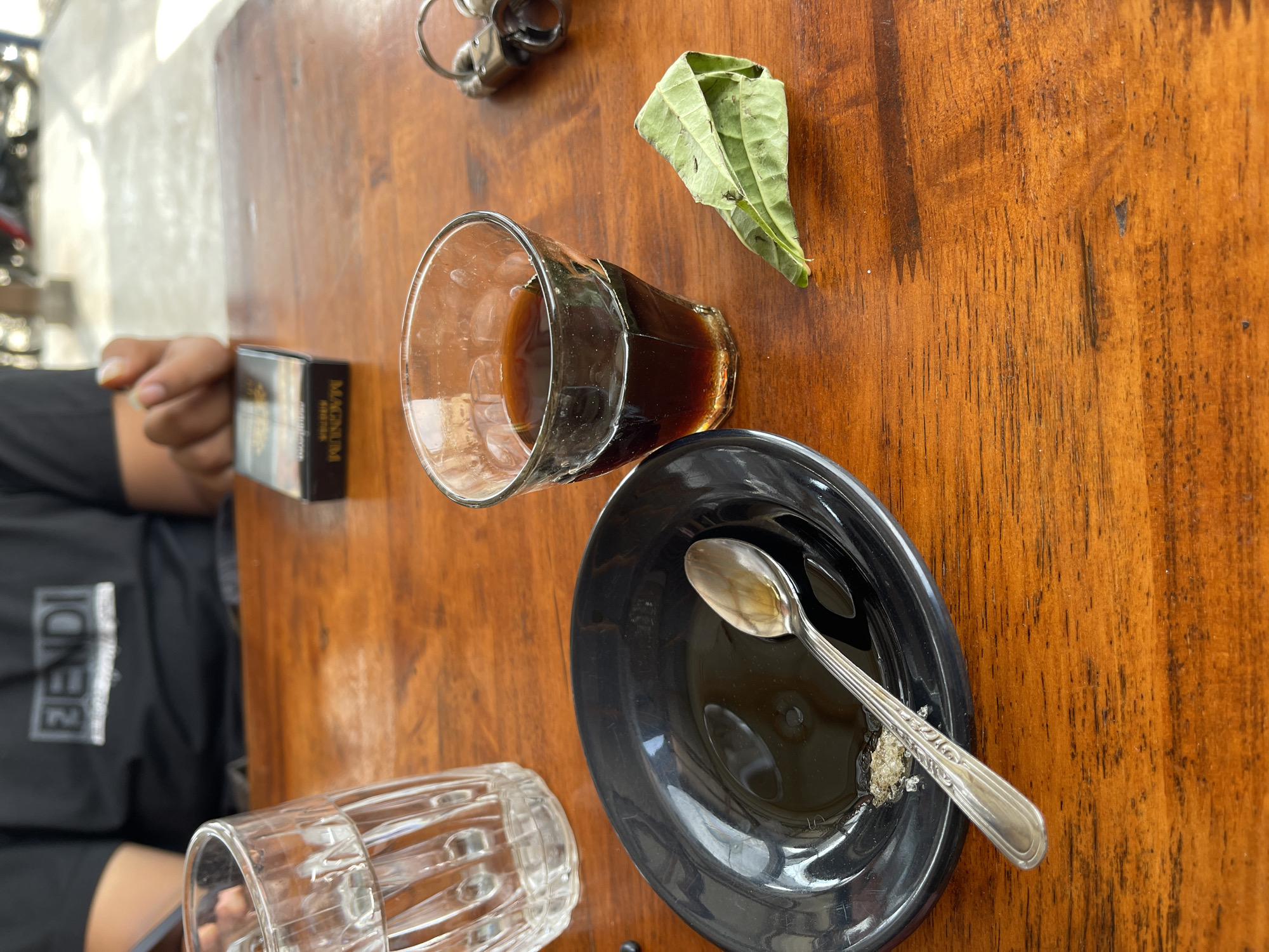 The image depicts a wooden table with several items on it, suggesting a casual setting, possibly an outdoor cafe or eatery, consistent with the general environment of Meunasahmee, Indonesia. A clear glass filled with a dark liquid, likely coffee or tea, is positioned on the right side of the table. To the left of this glass, a black plate contains a small amount of liquid and what appears to be coarse sugar crystals, with a silver spoon resting inside the plate. In the foreground, a folded green leaf lies on the table. A glass filled with water is visible in the upper left corner. In the background, a person wearing a black t-shirt with a "BENDI" logo is partially visible, and a pack of "MAGNUM" cigarettes is placed on the table near them. A set of keys with a silver fob is also present on the right side of the table. The lighting suggests it is daytime, with natural light illuminating the scene.