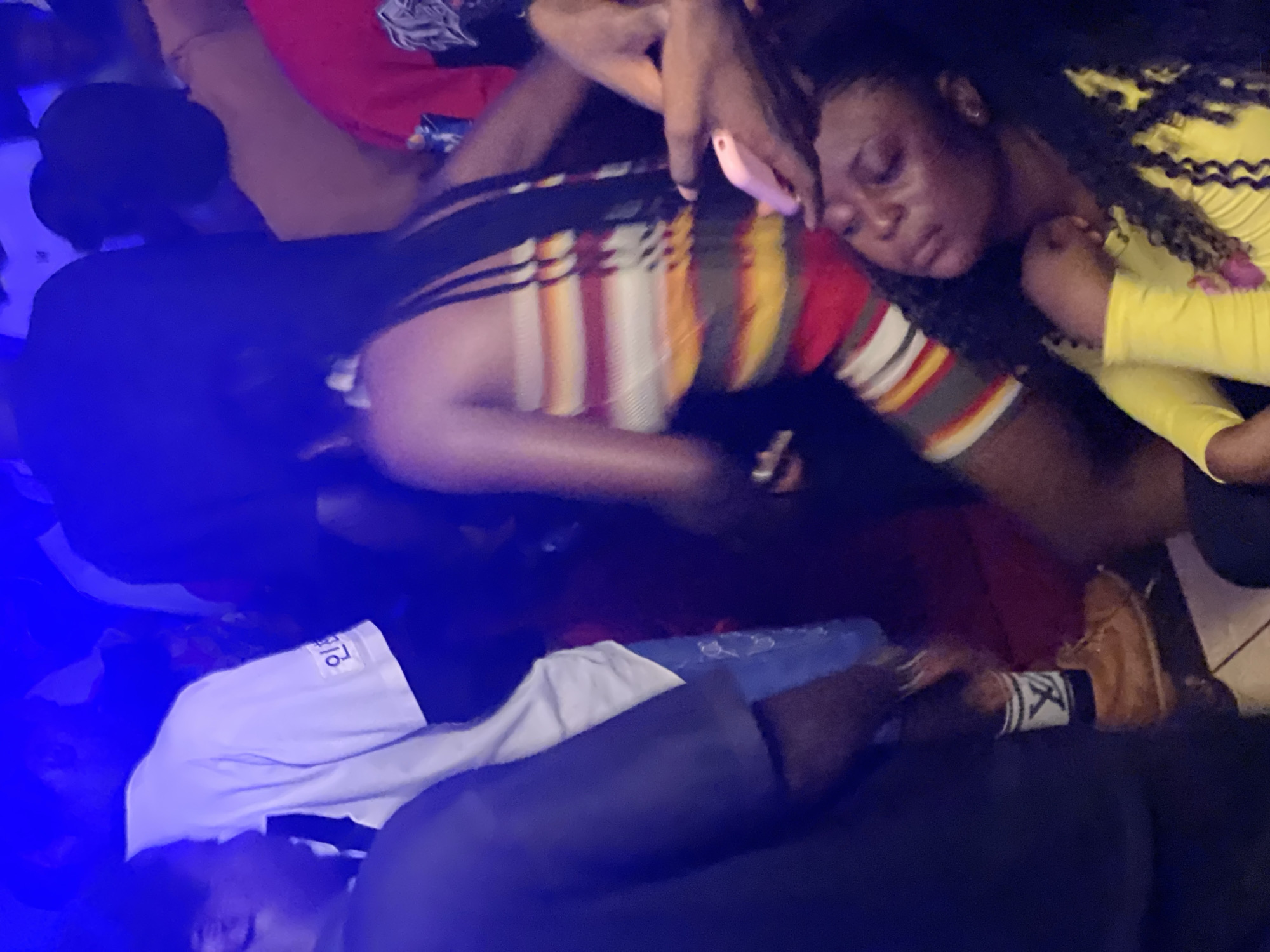 The image depicts a dimly lit indoor scene, likely a nightclub or party venue in Agwala, Nigeria, with vibrant blue and purple lighting dominating the atmosphere. In the foreground, a woman in a colorful striped tube top is seen dancing, her body angled away from the viewer. To her right, another woman, with her eyes closed and head tilted back, is wearing a bright yellow long-sleeved shirt and appears to be experiencing the music. Her left arm is bent, and her hand is near her chest, while her right hand is raised and holding a pink cell phone. In the background, a crowd of people are visible, most of them indistinct due to the low lighting and motion blur. One person in the middle ground wears a white t-shirt with Korean writing on it. The overall impression is one of energetic movement and a lively social gathering.