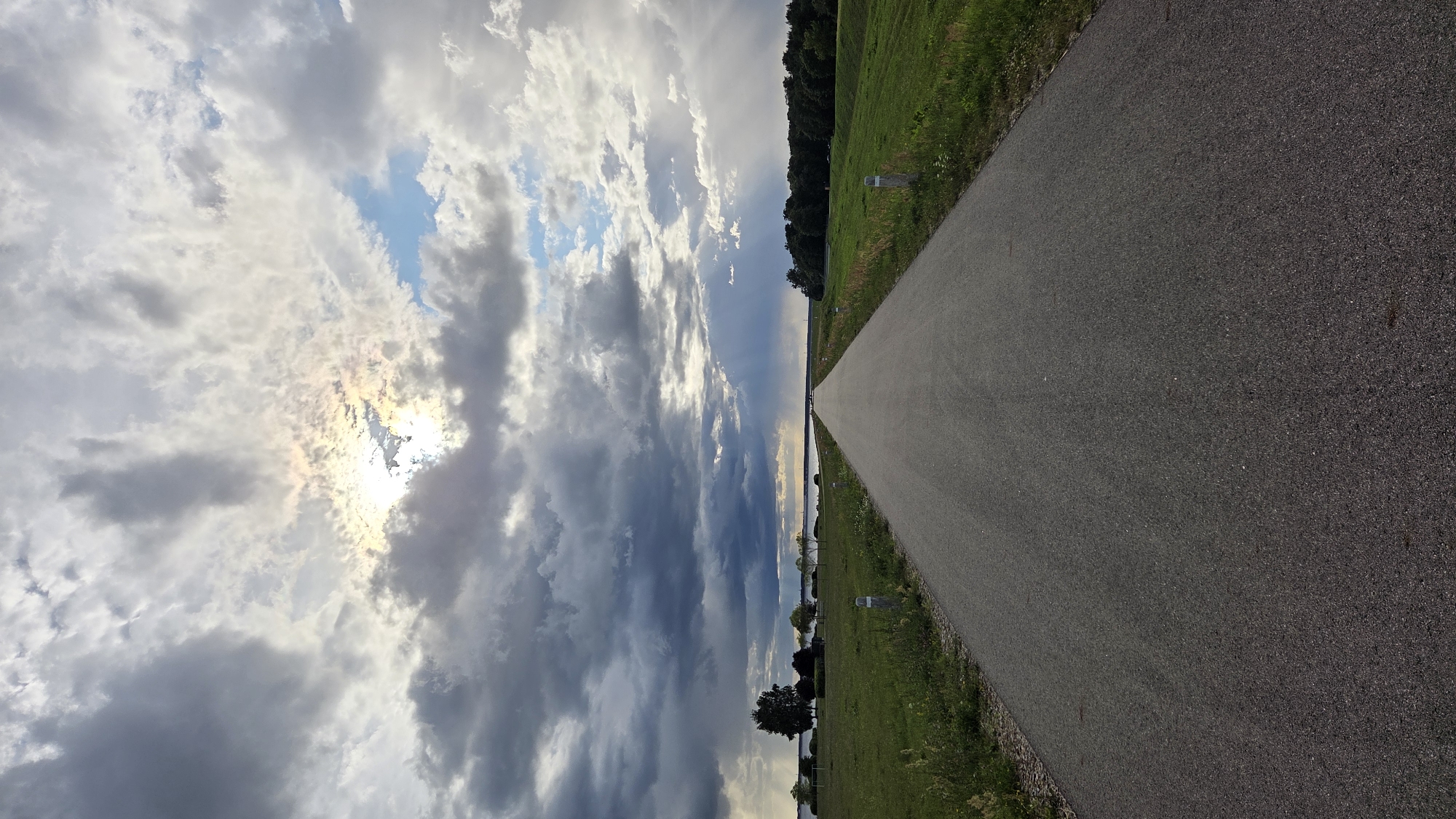 The image depicts a straight, paved road stretching into the distance, bordered by green grassy fields on both sides. The sky is dramatic, filled with a mix of dark, heavy clouds and patches of bright sunlight breaking through, creating a sense of an approaching or receding storm. In the distance, a body of water is visible, possibly a river or lake, with a line of trees along its far bank. The setting appears to be rural or semi-rural. While the exact location within Salaspils, Latvia is not explicitly identified, the general landscape is consistent with areas found in the vicinity of the Daugava River, which flows through or near Salaspils. There are no people or active events captured in the scene.