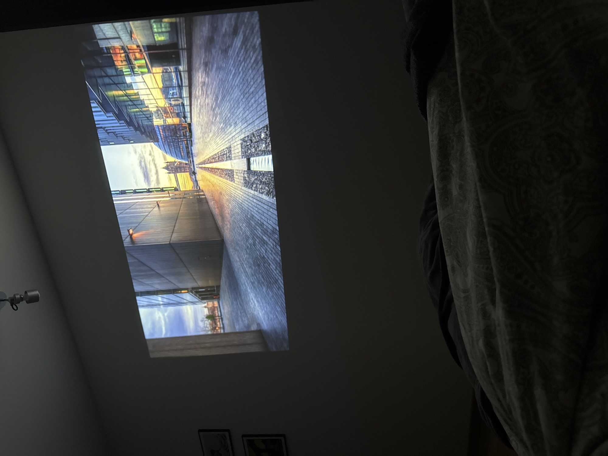 A photograph taken indoors from a low angle displays an image projected onto a wall, likely from a projector in a darkened room. The projected image depicts a contemporary urban environment, specifically a view of the pedestrian bridge in Warsaw, Poland, at sunrise. The architecture is modern, with buildings featuring glass and metal facades. The pedestrian bridge is constructed from brick. To the right of the projected image, partially obscured by shadows, is a draped fabric. The camera is positioned close to the fabric. Additional details include a wall fixture and partially visible framed art on the wall below the projected image.