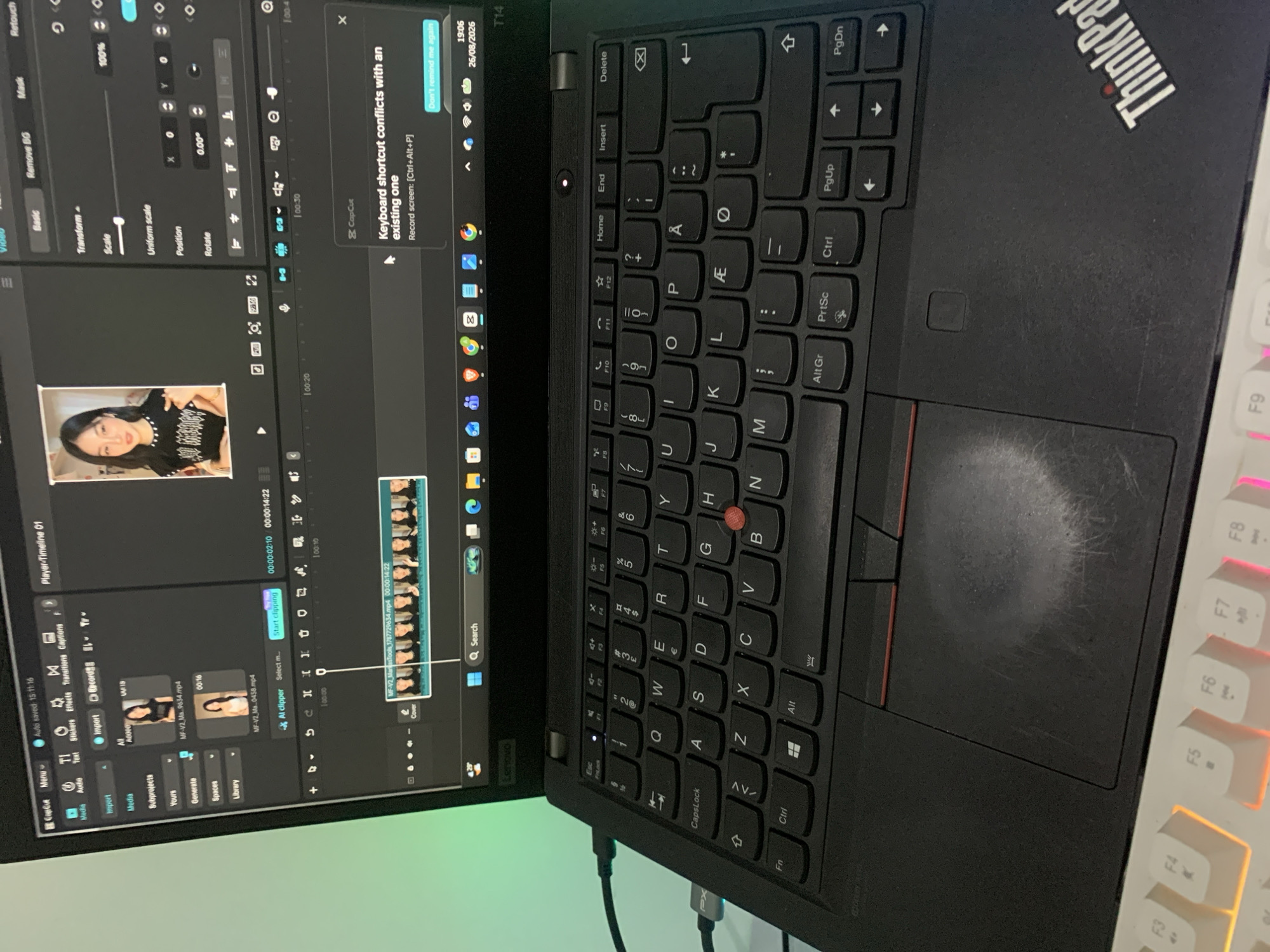 The image shows a close-up of a Lenovo ThinkPad laptop, open and displaying the CapCut video editing software. The screen shows a video project with a timeline and various editing tools. There are two video clips, identified as "MF-V2_Ma_9634.mp4" and "MF-V2_Ma_0438.mp4", loaded into the project. A pop-up window indicates a "Keyboard shortcut conflicts with an existing one". The laptop is on a desk, and a portion of a white keyboard with RGB lighting is visible in the foreground. The background is out of focus but appears to be an indoor environment with a greenish hue. The timestamp on the laptop screen shows "19:06 26/08/2026". The location of Dukuh Mojo, Indonesia is not visible in the image.