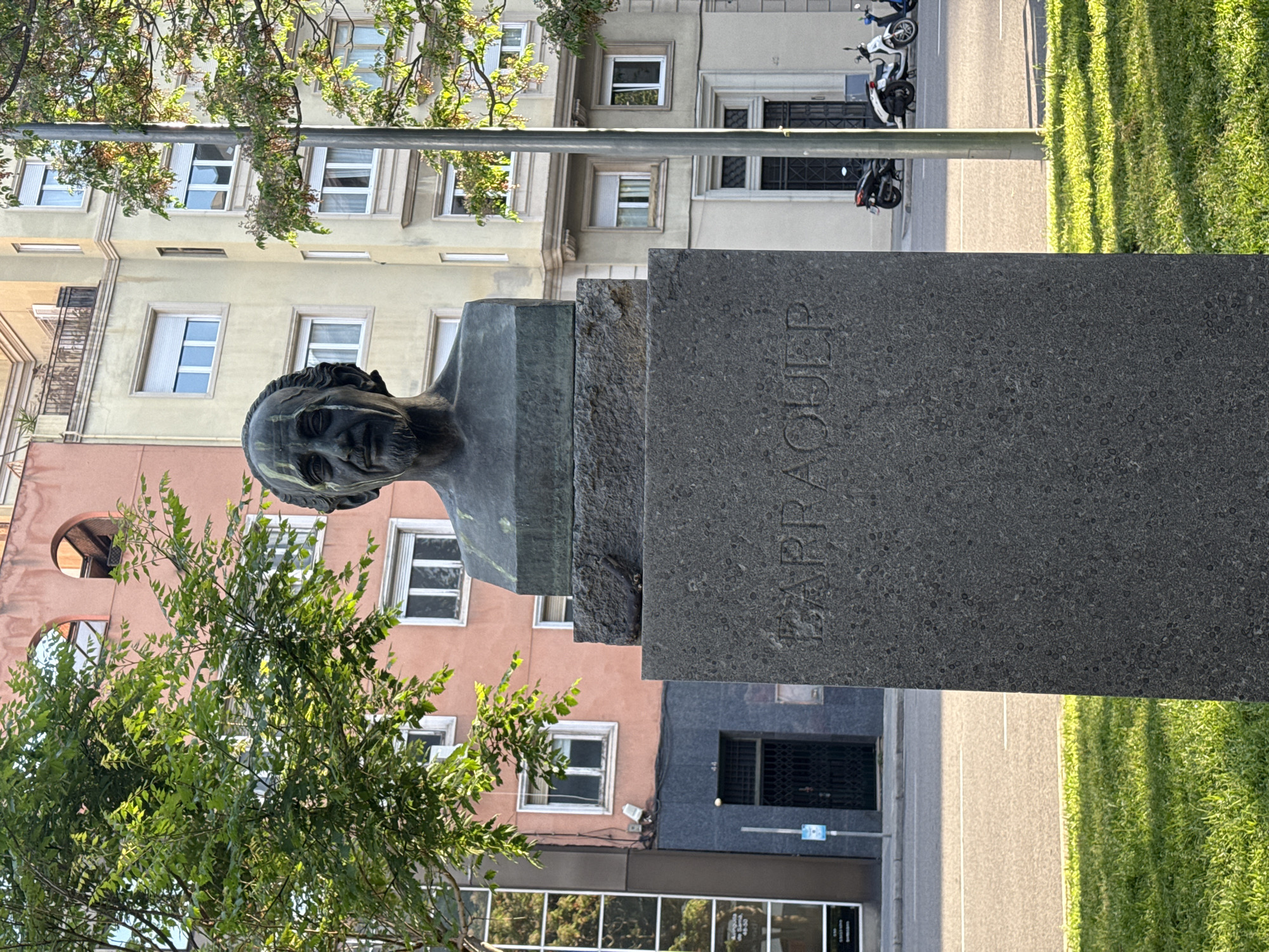 The image shows a bronze bust of a man atop a stone pedestal, situated outdoors in what appears to be a park or public area. The name "BARRAQUER" is engraved vertically on the pedestal. A tree is partially obscuring the bust from the viewer's perspective. In the background are buildings of varied colors and architectural styles, with windows and balconies visible. A street runs alongside the monument, with parked motorcycles on one side and a strip of green grass or foliage on the other. The overall impression is of a sunny, urban setting with a monument dedicated to someone named Barraquer. I believe it is on Avinguda Diagonal in Barcelona, Spain.