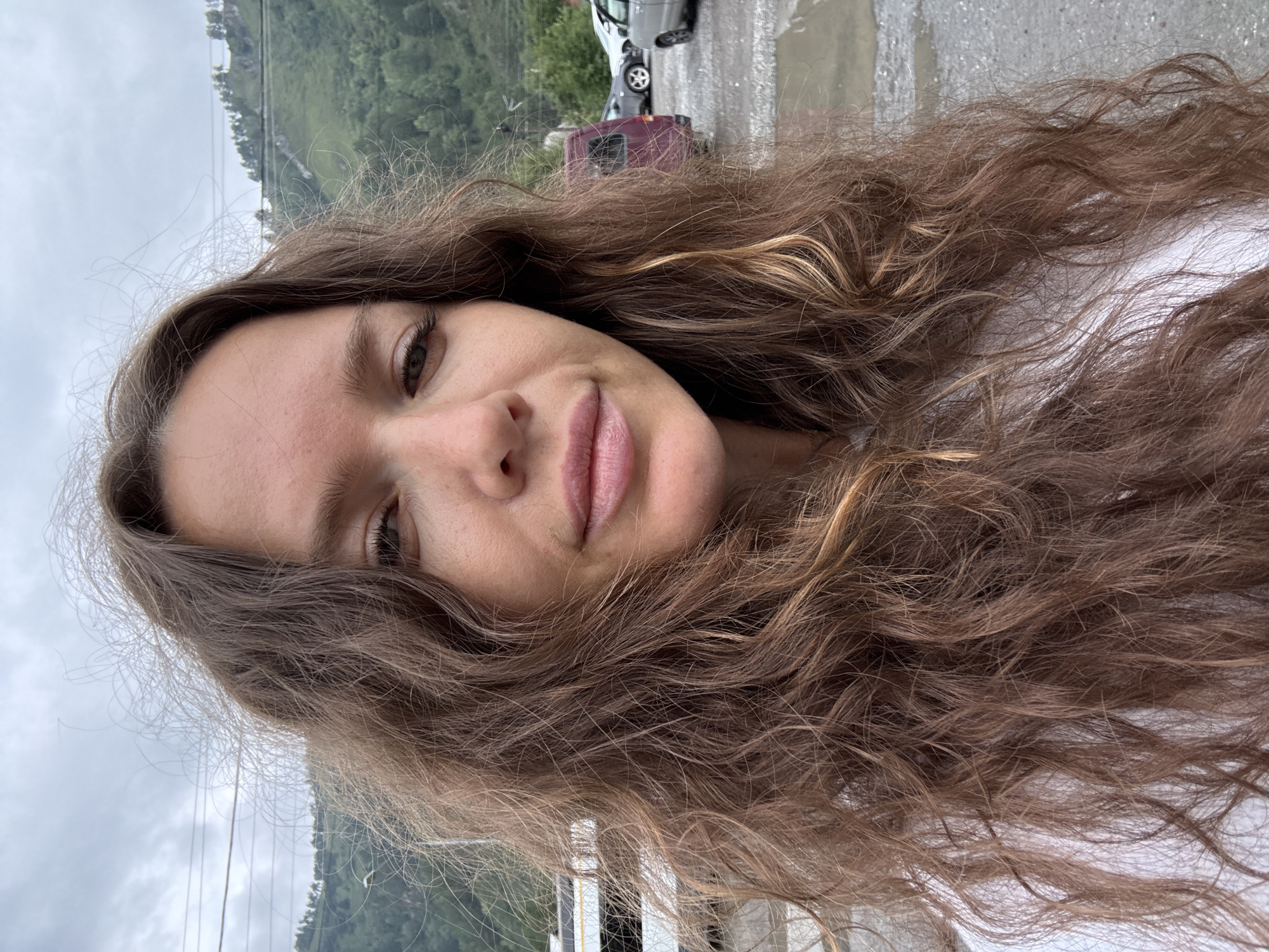 This is a selfie of a woman with long, wavy brown hair. She is looking directly at the camera with a slight smile. The background appears to be an outdoor setting, possibly a parking lot or a street, with mountains and trees visible in the distance. There are power lines overhead and a few vehicles are parked in the background. The sky is overcast. The woman is wearing a white top. The image was taken in Manzherok, Russia.