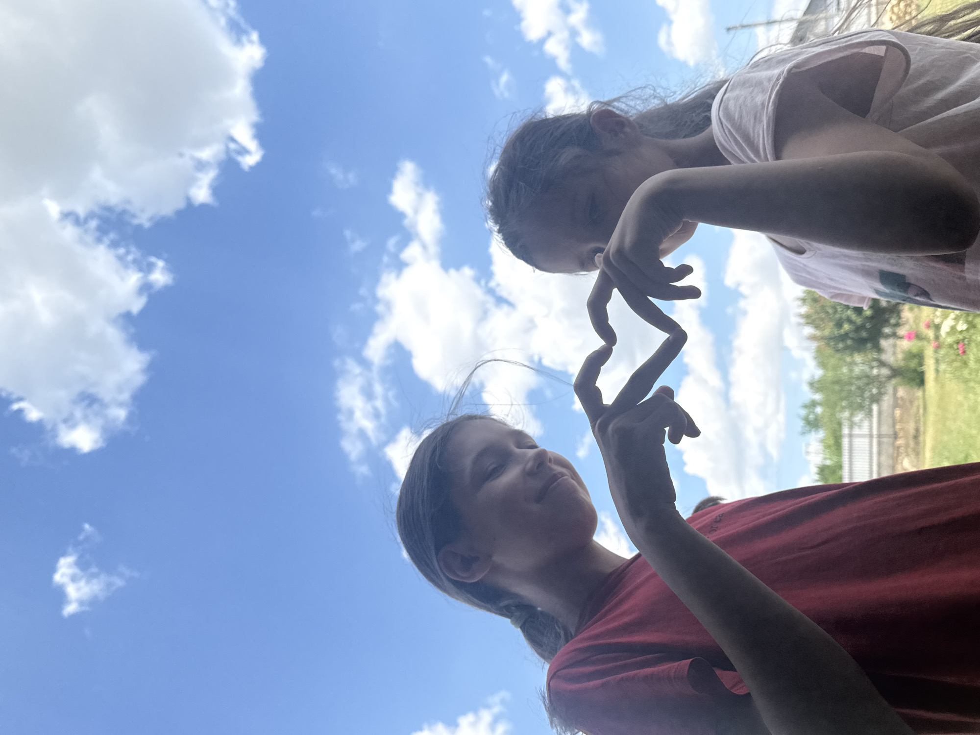 Two young girls are silhouetted against a bright blue sky with scattered white clouds. They are standing outdoors, facing each other, and forming a heart shape with their hands in the center of the frame. The girl on the left is wearing a red t-shirt, and the girl on the right is wearing a light-colored t-shirt. In the background, out of focus, is a green grassy area, a white fence, and some trees. The lighting suggests it is daytime.