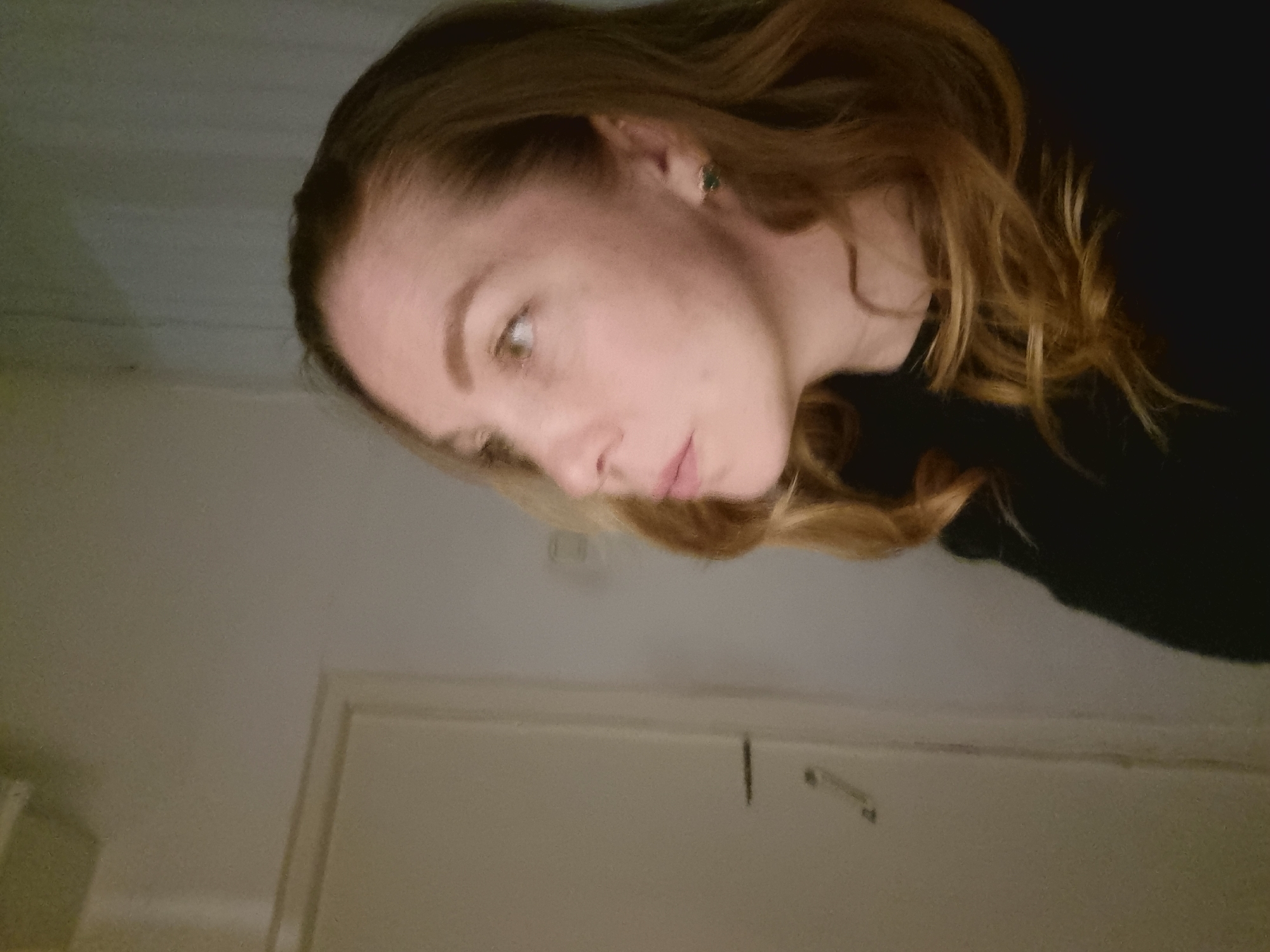 The image depicts a woman indoors. She has light skin, light brown to blonde hair, and is wearing a dark top. The background shows a room with a white or light-colored wall and a door. The lighting is dim. The location in the city of Kemul, Russia is not determinable from this image.