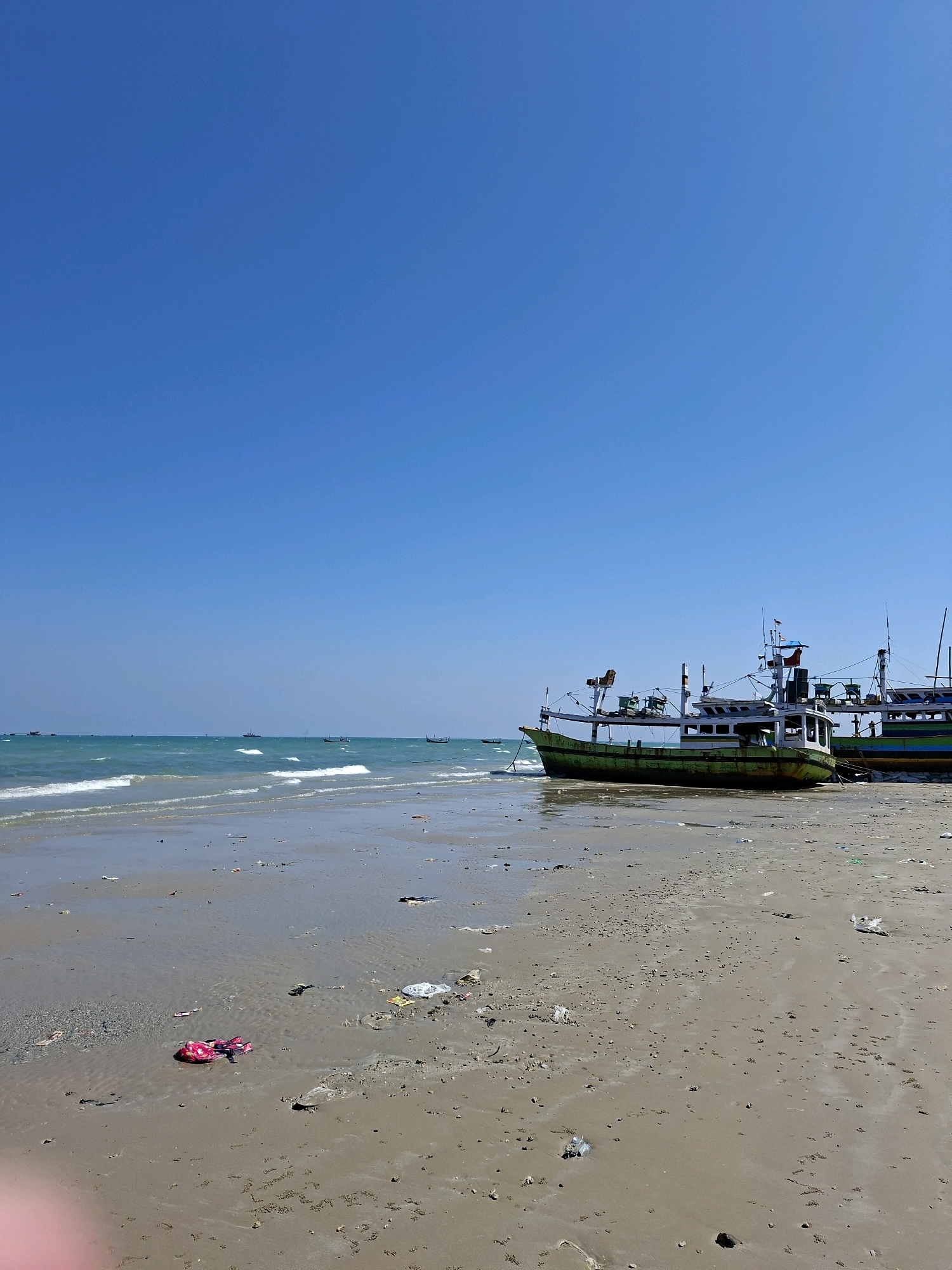 The image shows a beach scene at Camplong, Indonesia. The sky is clear blue. Several fishing boats are anchored on the sand and in the shallow water. The beach is sandy and appears to have some debris scattered across it. The water is turquoise and there are small waves. The scene depicts a typical day at a coastal location.