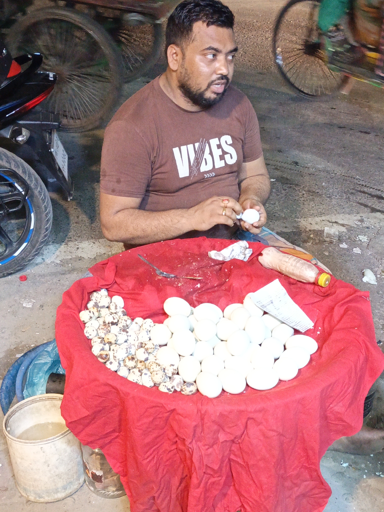 In an outdoor setting in Unknown, Unknown, a man with a beard and a brown "VIBES" T-shirt sits behind a red-clothed table filled with eggs. To the left are speckled quail eggs, and to the right, chicken eggs. The man is peeling an egg. A spoon, a condiment bottle, and paper sit on the table. A motorcycle is parked to the left of the table. A bicycle wheel is visible in the background. A bucket and other items are near the bottom of the frame.