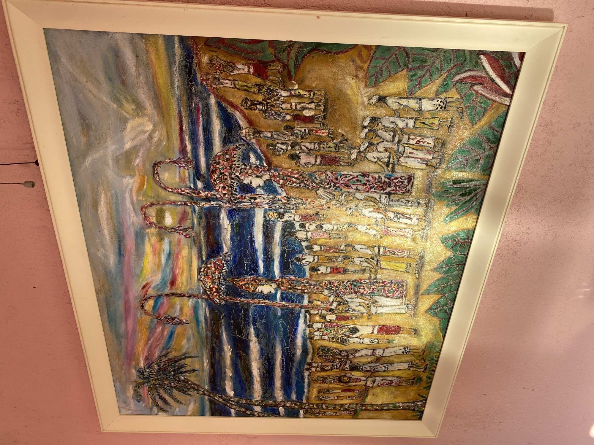 A framed, horizontal oil painting on canvas depicts a vibrant scene of a procession or festival on a beach. The sky is a riot of colorful, impressionistic streaks of pink, orange, yellow, and blue, suggesting a sunset or sunrise over a choppy, dark blue sea. Two palm trees stand on the left, their fronds rendered in textured brushstrokes.
In the foreground and midground, a large gathering of people, primarily depicted in simple, stylized figures, are engaged in a procession. Many figures carry objects on their heads, possibly baskets or offerings, while others appear to be playing musical instruments. Two central figures are elaborately dressed and adorned with ornate headdresses and patterned garments. One of these figures holds a large, decorative umbrella. The figures are standing on a golden-hued beach, with hints of green foliage and red flowers in the lower right quadrant. The painting's surface shows a network of fine cracks, characteristic of aged oil paint. The artwork is housed in a plain, cream-colored frame and is displayed on a pink, textured wall.