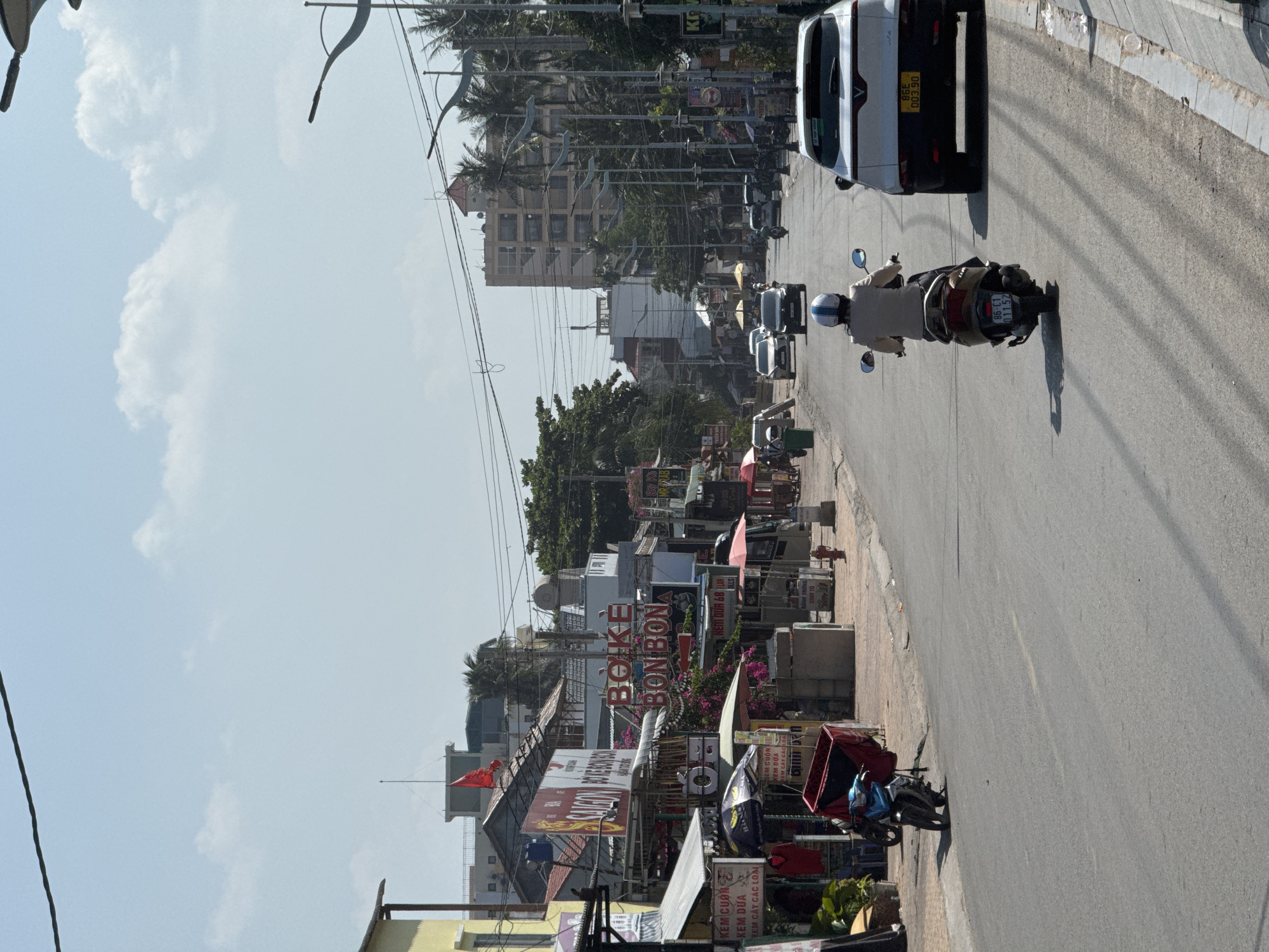 The image depicts a street scene in Mũi Né, Vietnam, under a clear sky with scattered clouds. The perspective is from a street-level viewpoint, looking down a paved road lined with shops and buildings on both sides.
On the left side of the road, several businesses are visible, including one with a prominent sign that reads "BOKE BON BON" in red letters. Other signs are in Vietnamese. There are awnings, umbrellas, and items displayed outside the shops, suggesting a commercial area. A parked blue and black motorcycle is visible near the sidewalk.
The road itself is a two-lane street with a white line dividing the lanes. Several vehicles are present: a white SUV is in the right lane, and a scooter with a rider wearing a light-colored jacket and a white helmet is in the left lane, slightly ahead of the SUV. Further down the road, other cars and motorcycles can be seen.
Palm trees and other foliage line the right side of the street, interspersed with streetlights and more businesses. In the background, several multi-story buildings rise above the single-story shops. The architecture appears to be a mix of modern and older styles.
The overall atmosphere is that of a bustling, sunlit street in a coastal town. The presence of shops, vehicles, and pedestrians indicates normal daily activity. The image captures a typical street view, highlighting the blend of commercial life and tropical vegetation characteristic of Mũi Né.