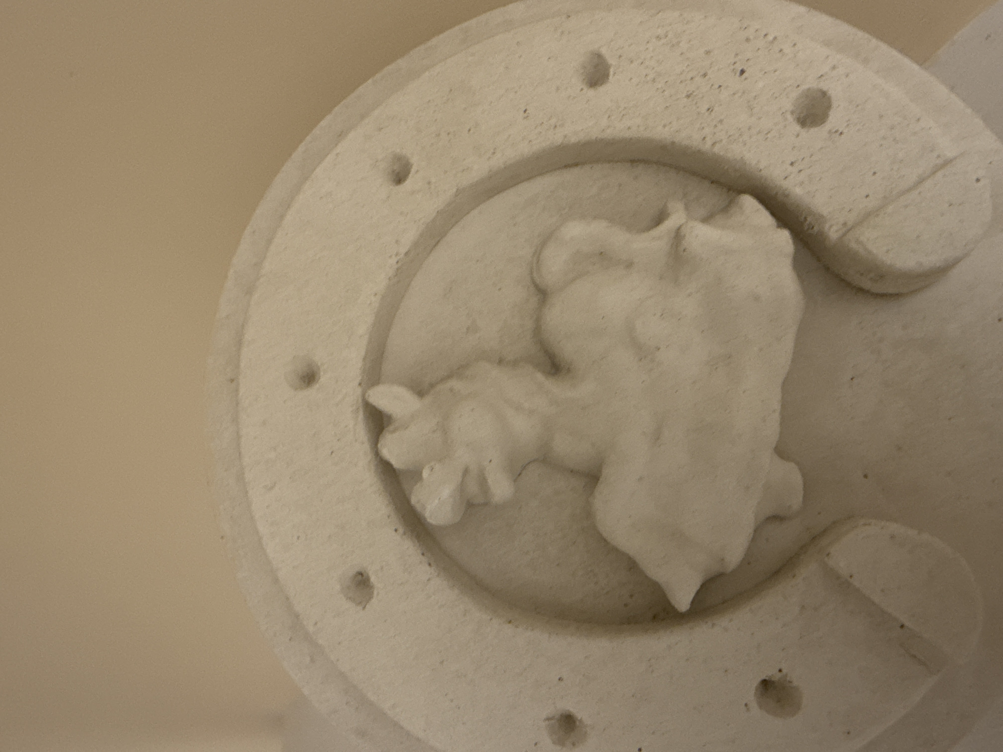 A close-up photograph depicts a stone horseshoe with a raised, abstract figure inside. The horseshoe has a series of small holes around its perimeter. The entire sculpture is a light-colored stone. Based on its appearance and subject matter, this could be a decorative architectural element. No people, specific actions, or interactions are visible in the image.
While I do not know the specific location of this particular object in Votkinsk, Russia, the city is known as the birthplace of the composer Pyotr Ilyich Tchaikovsky, and therefore it is possible that this horseshoe is part of a monument or architectural feature dedicated to him or to another cultural aspect of the city. Without more information, it is impossible to be certain of the object's precise origin or function.