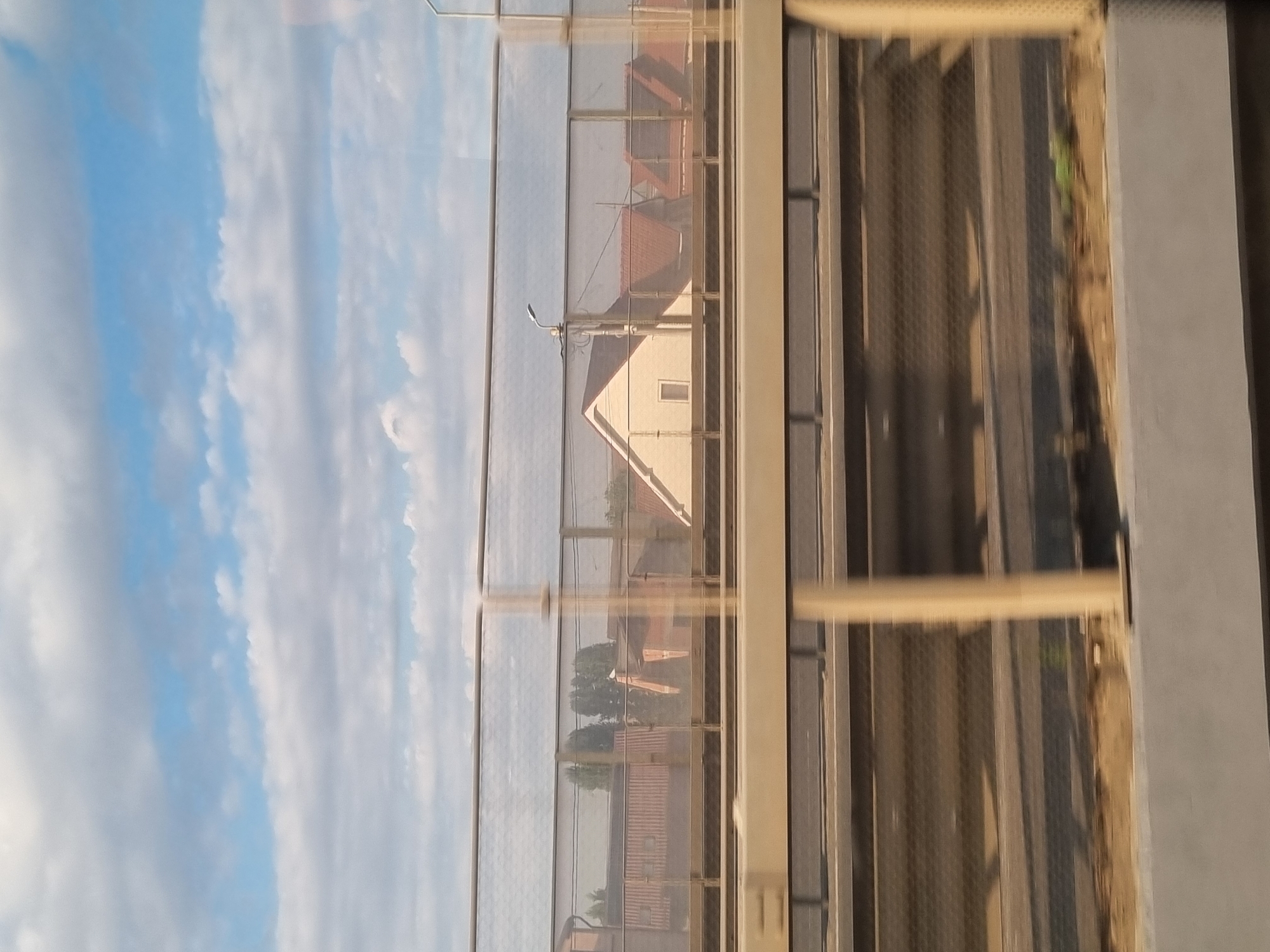 The image depicts a view through a protective barrier, likely a sound barrier on a bridge or elevated road, overlooking a residential area with houses. The sky is partly cloudy with blue patches. Below the barrier, there appears to be a road or railway with blurred motion indicating movement. The houses visible have red-tiled roofs and light-colored walls. A street lamp is visible on a pole in the distance. The overall scene is a snapshot of a suburban or semi-urban landscape as seen from a moving vehicle or elevated position. The location is identified as Oradea, Romania.