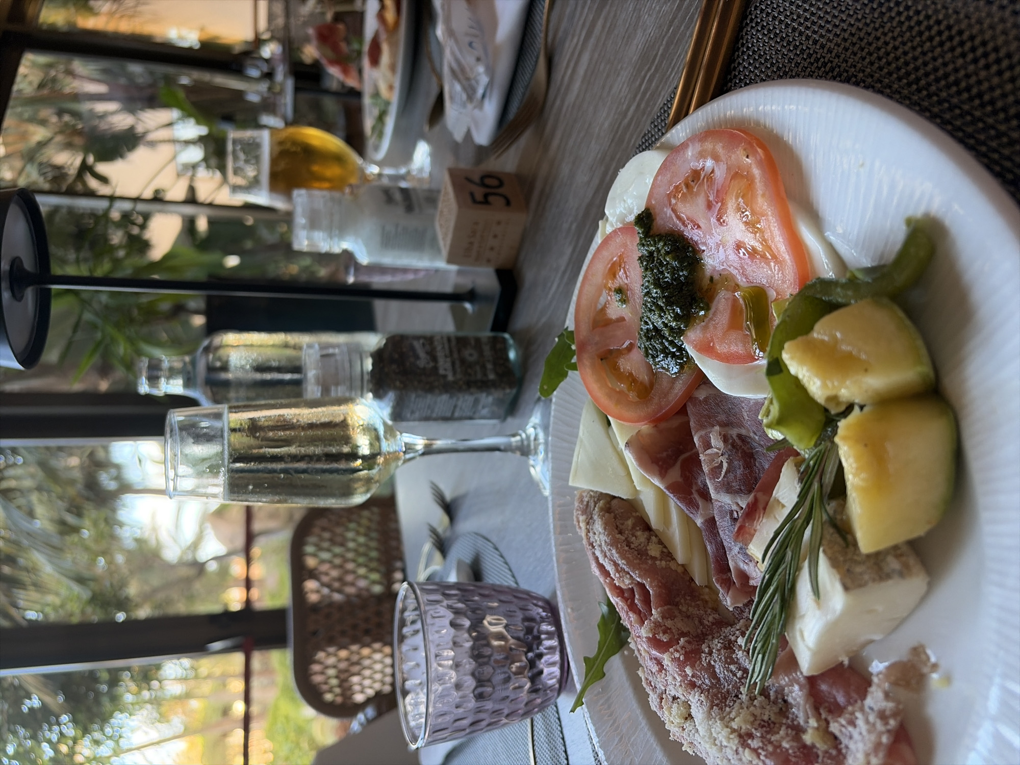The image presents a close-up, angled view of a dining table set with food and drinks. The primary subject is a white plate laden with an assortment of appetizers. This includes slices of red tomatoes paired with fresh mozzarella and a dollop of green pesto, alongside various cured meats, some sprinkled with white cheese, and different types of cheeses, including a soft white one and others with rinds. A sprig of rosemary and some green peppers garnish the plate, with arugula visible underneath.
Beside the plate, a wine glass holds white wine, and a purple, textured glass sits on a patterned placemat. Two clear shakers or bottles are also present on the wooden table; one contains black peppercorns, while the contents of the other are indistinct. A small wooden block with the number "56" and the text "Vila Sira" is visible.
The background is softly blurred, showing reflections of green foliage and natural light, indicating an outdoor or semi-outdoor setting with pleasant daytime or late afternoon lighting. The scene suggests a relaxed meal, possibly at a restaurant or resort in Antigua, Spain.