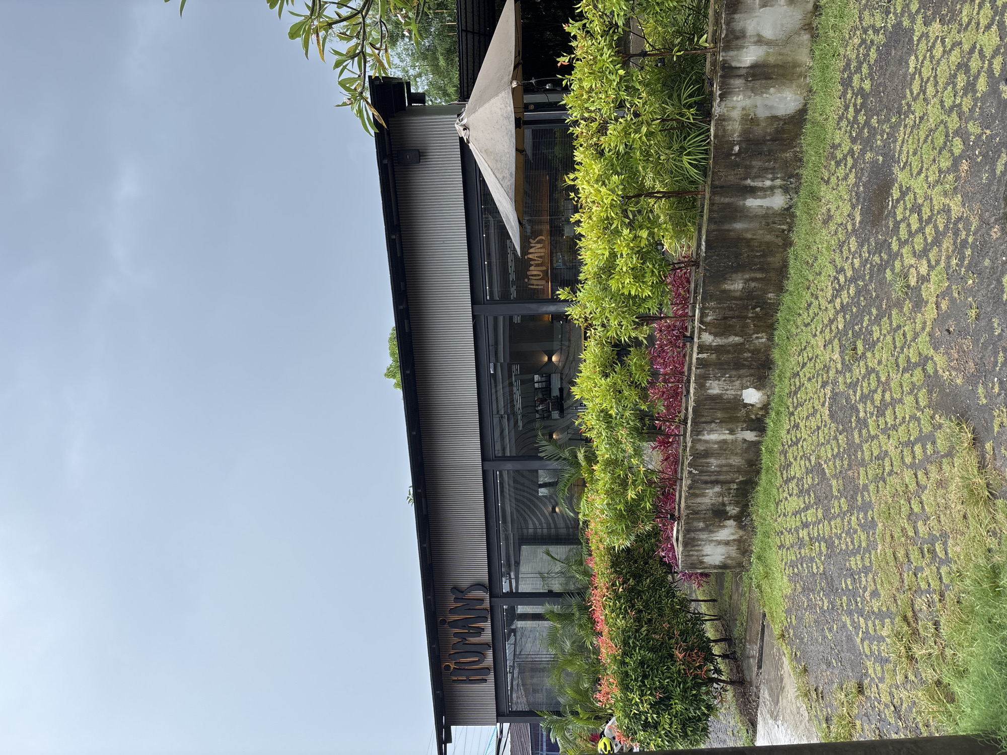 The image depicts the exterior of a modern building, likely a commercial establishment such as a cafe or restaurant, located in Kuta Selatan, Indonesia. The building features a contemporary design with a dark, corrugated metal upper facade and large, expansive glass windows on the lower level, some of which show internal blinds. A prominent, stylized sign on the glass facade spells out "HUMANS". An off-white or light-colored awning extends from the building, providing shade over a section of the entrance area.
The building is surrounded by lush greenery, indicative of a tropical or subtropical climate. A vibrant arrangement of bushes with both green and reddish foliage runs along the front of the building. To the right, these plants are bordered by a low, weathered concrete wall, showing signs of discoloration and staining. Beyond this wall, the ground is a mix of dark, damp-looking earth and sparse, patchy green grass. Additional green foliage, including larger plants and small trees, is visible further in the foreground and background, contributing to the natural ambiance.
The sky above is a clear, light blue with subtle, diffused light, suggesting a daytime scene, possibly under a slightly overcast sky or during the softer hours of morning or late afternoon. No individuals or specific activities are visible, presenting a serene and still architectural landscape.
