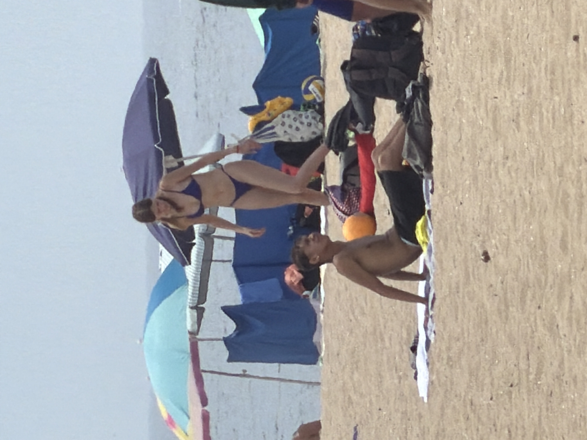 The image depicts a beach scene in Espinho, Portugal, with several individuals and beach accessories. In the foreground, a young man with dark hair is seated on a white towel on the sand, facing to the right and looking away from the camera. He is shirtless and wearing dark shorts. Behind him, a young woman in a blue bikini is standing and looking down. To her left, a large purple umbrella provides shade. Further back, there are blue tents and other beach umbrellas visible. Various items such as bags, a volleyball, and possibly clothing are scattered on the sand. The sea and sky form the background.