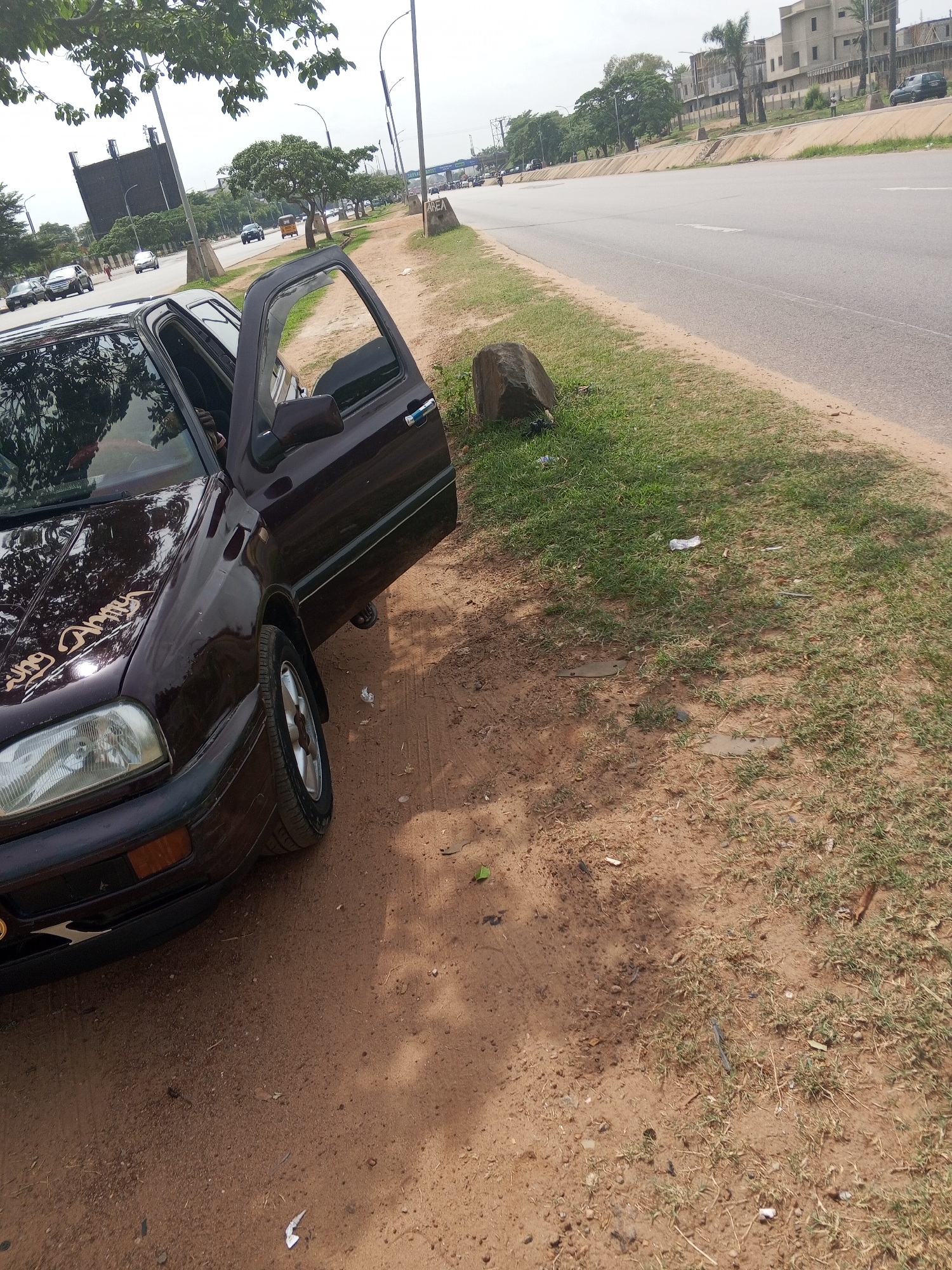 The image shows a dark burgundy Volkswagen Golf parked on a dirt shoulder next to a paved road. The driver's side door is open. The car has a sticker on the hood, partially obscured by reflections. The ground is a mix of red dirt, grass, and scattered litter. The paved road is gray with a painted white line. In the background, a wide road with light traffic is visible, flanked by buildings and trees. The scene is likely in Abuja, Nigeria, characterized by the architecture and vegetation of the city.