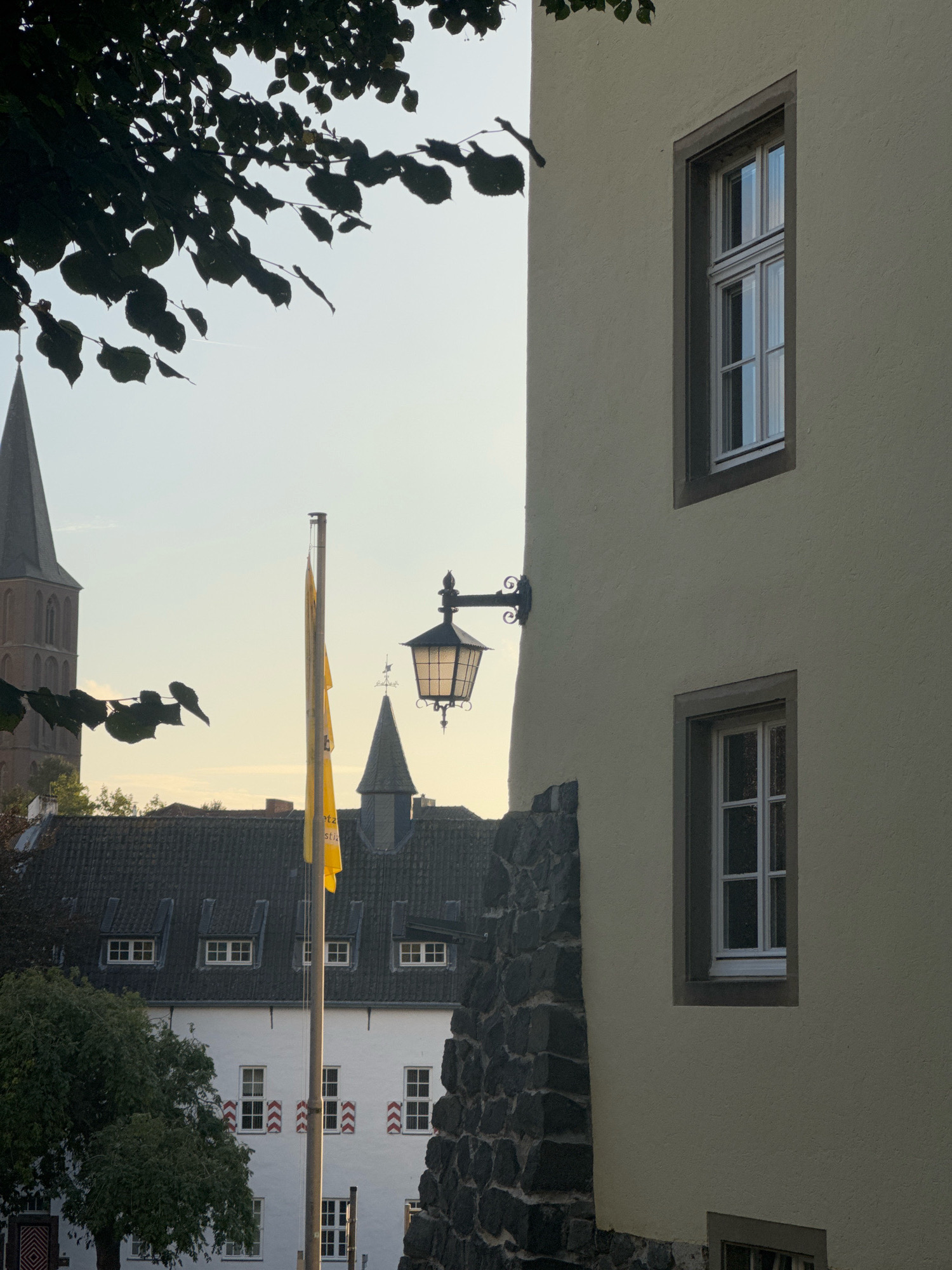 The photograph shows a view of Cleves, Germany. The foreground features the side of a building with a pale yellow, textured facade. Two rectangular windows with white frames are visible on this facade. The lower part of the building's exterior is a rough stone wall. To the left of this building, a yellow flag is mounted on a pole. Behind the flag, a dark-tiled roof with several dormer windows is visible. Beyond the roof, a church steeple with a dark gray cap rises into the sky. To the far left, the upper portion of another, taller church steeple is visible, also with a dark gray cap. A wrought-iron lamppost with a hexagonal glass lantern is attached to the building's facade. The sky is a pale, hazy blue. Dark green leaves and branches from a tree are silhouetted against the sky in the upper left corner of the image. The scene appears to be set in the late afternoon or early evening, given the soft lighting.