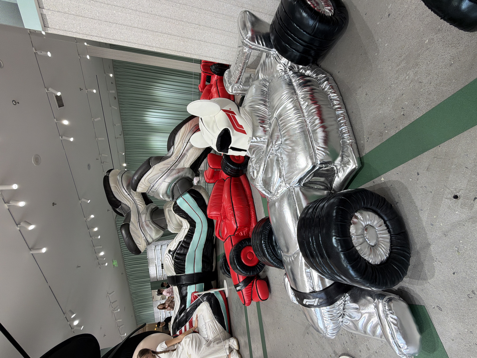 The image shows an indoor art installation featuring inflated, oversized sculptures of racing-themed objects. The sculptures include a Formula One race car, racing gloves, racing boots, and a helmet. The race car is primarily silver with black tires and details. The gloves are red and white, while the boots are various colors, including silver, black, and light blue. The installation appears to be in a gallery or exhibition space, with a concrete floor and overhead lighting. People are visible in the background, observing and interacting with the artwork. The image was taken in Jing'an District, China.