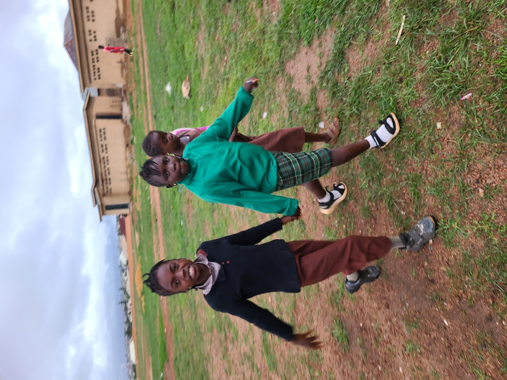 The image shows three children, likely girls, outdoors on what appears to be a school ground or open field in Jos, Nigeria. The photograph is oriented vertically, rotated 90 degrees clockwise from its natural horizontal perspective.
Two of the children in the foreground are clearly visible and smiling, appearing cheerful. The child on the left wears a dark blue long-sleeved top, a light-colored collared shirt with a plaid pattern, brown trousers, and dark closed-toe shoes. Her arms are slightly outstretched, and her braids are pulled back. The child in the center wears a bright green long-sleeved top, plaid shorts, and black and white open-toed sandals. Her arms are also slightly extended. A third child is partially visible behind the central figure, wearing a pink top and a brown skirt.
The ground is a mixture of reddish-brown dirt and patches of green grass, suggesting a well-used outdoor area. In the background, several light-colored, multi-story buildings with multiple small windows are visible, one with a reddish-brown roof. The architecture appears functional and simple. The sky is overcast and hazy, suggesting a cloudy daytime. A distant landscape with more structures is faintly visible under the cloudy sky. No text is visible in the image. The children appear to be in a playful mood, possibly posing for the camera.