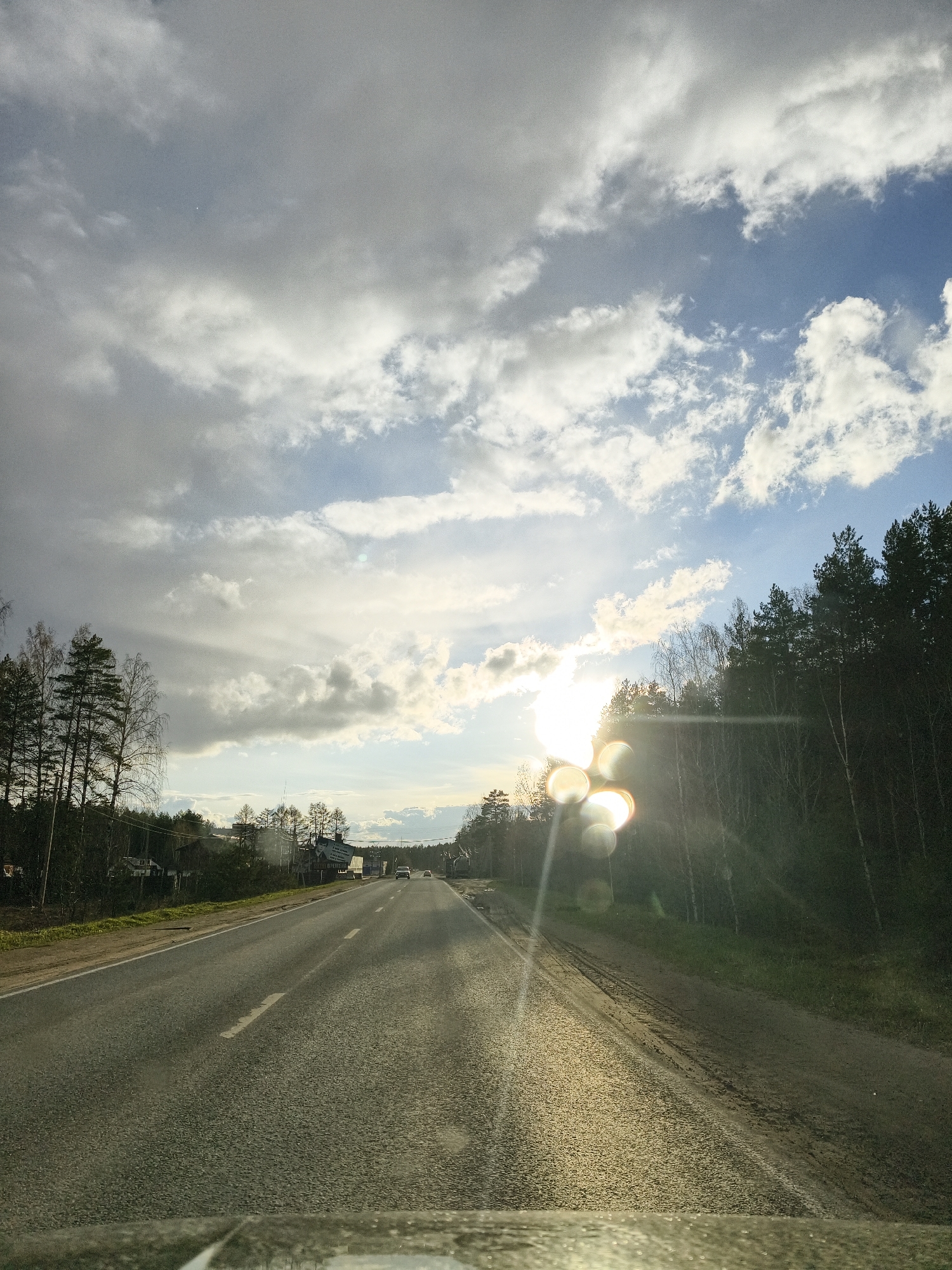 The media file presents a straight asphalt road, viewed from the interior of a vehicle. The road, featuring a faint white dashed center line, extends into the horizon. On both sides, dense vegetation comprised of coniferous trees (pines) and bare deciduous trees (likely birches) frames the path, suggesting a late autumn or early spring season.
On the left side of the road, nestled behind the trees, are glimpses of distant buildings. On the right, further along, a large sign or billboard with unreadable Cyrillic text is visible, partially obscured by trees.
The sky is a prominent feature, showing a mix of bright white and grey clouds with patches of clear blue. The sun is low in the sky, positioned on the right side, shining directly towards the viewer. This creates significant lens flares and a bright, golden reflection across the road surface and the vehicle's windshield at the bottom of the frame. The strong sunlight bathes parts of the road and the tree line in a warm glow.
A few vehicles can be seen in the far distance on the road, indicating ongoing traffic. The time of day appears to be late afternoon or early evening, characterized by the sun's low angle. The overall setting is a semi-rural or forested landscape, consistent with the provided location context of Noviy, Russia. The bottom edge of the image reveals reflections from the car's dashboard or windshield, confirming the viewpoint from inside a moving vehicle.